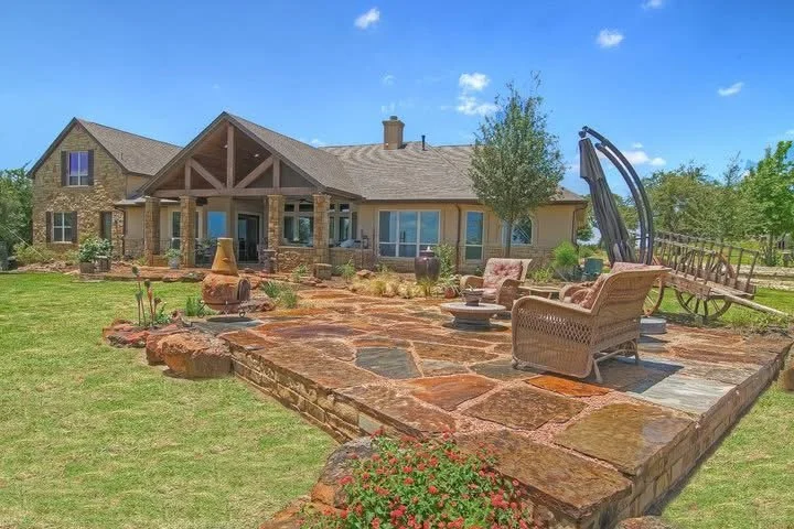 View of a large backyard patio with outdoor furniture, decorative pots, and a mounted wagon wheel, adjacent to a house with a porch and a landscaped lawn.