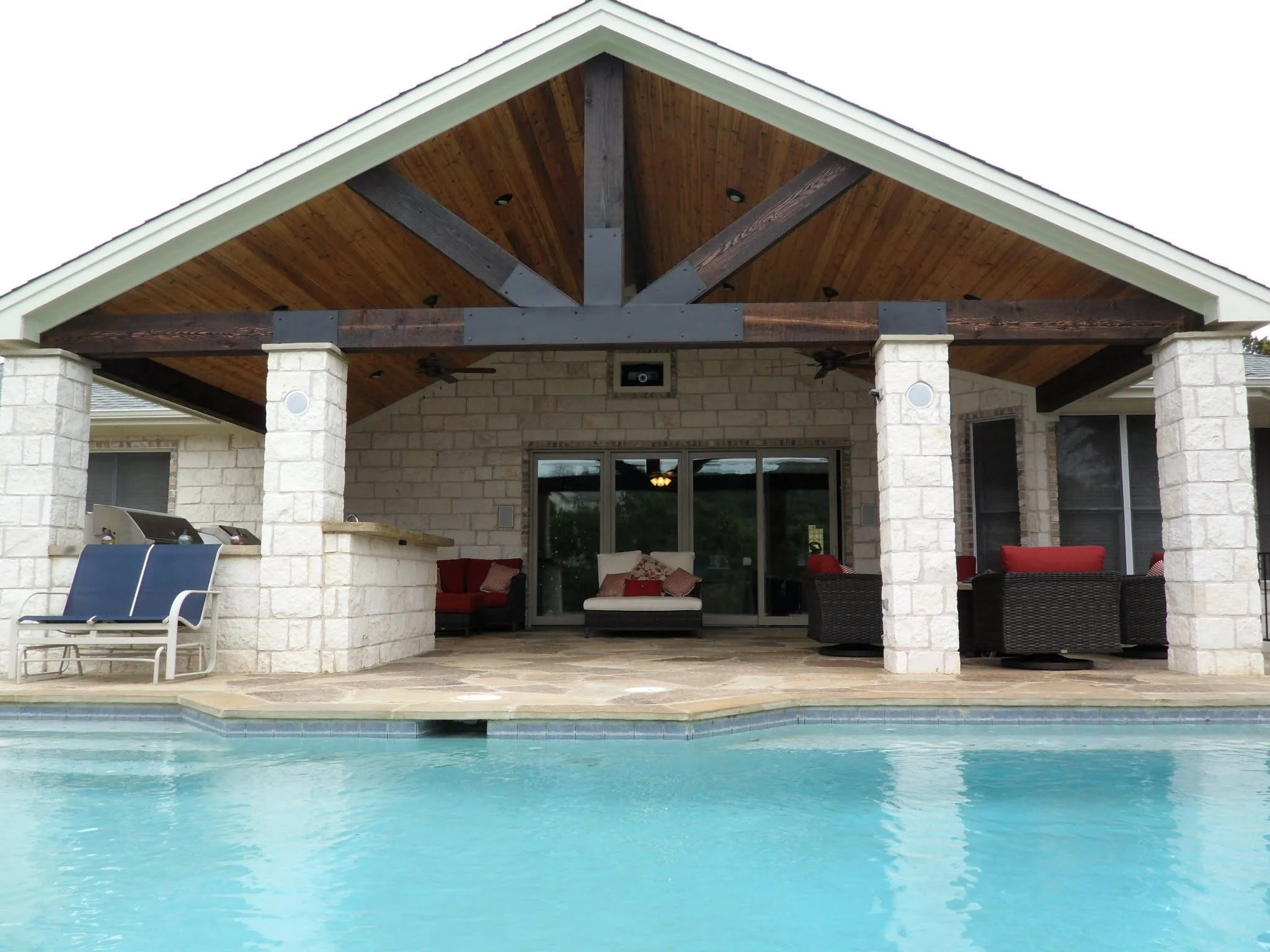 Residential backyard with clear swimming pool, covered patio area featuring outdoor furniture, brick columns, and a wooden gabled ceiling, with glass sliding doors leading inside.