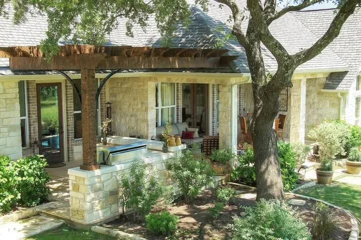 Front porch and garden of a house with stone walls, a large tree, potted plants, and outdoor furniture.