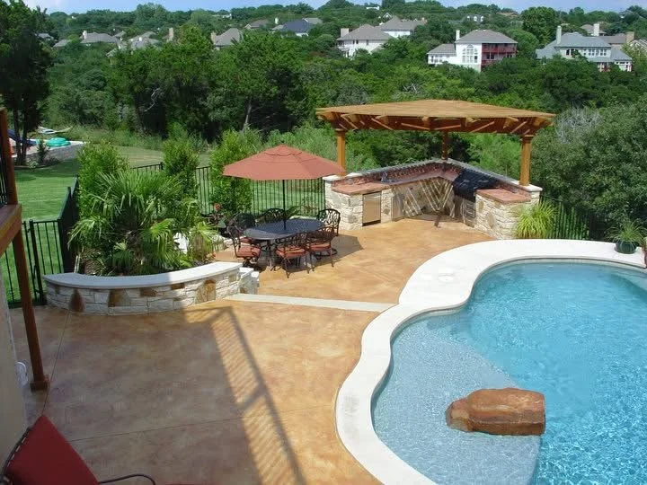 View of a backyard pool area with a curved swimming pool, a stone patio, a patio table with chairs and an umbrella, a shaded outdoor kitchen area with a wooden roof, and a lush green landscape with houses in the background.
