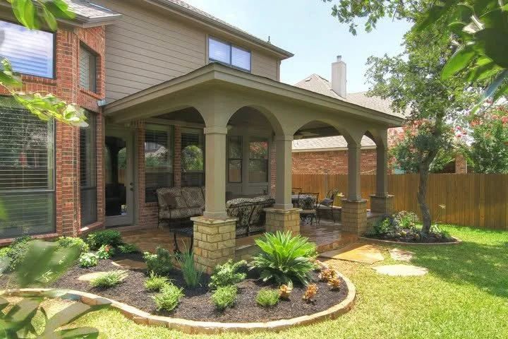 Backyard patio with covered seating area, brick pillars, outdoor furniture, surrounded by a well-maintained garden and wooden fence.
