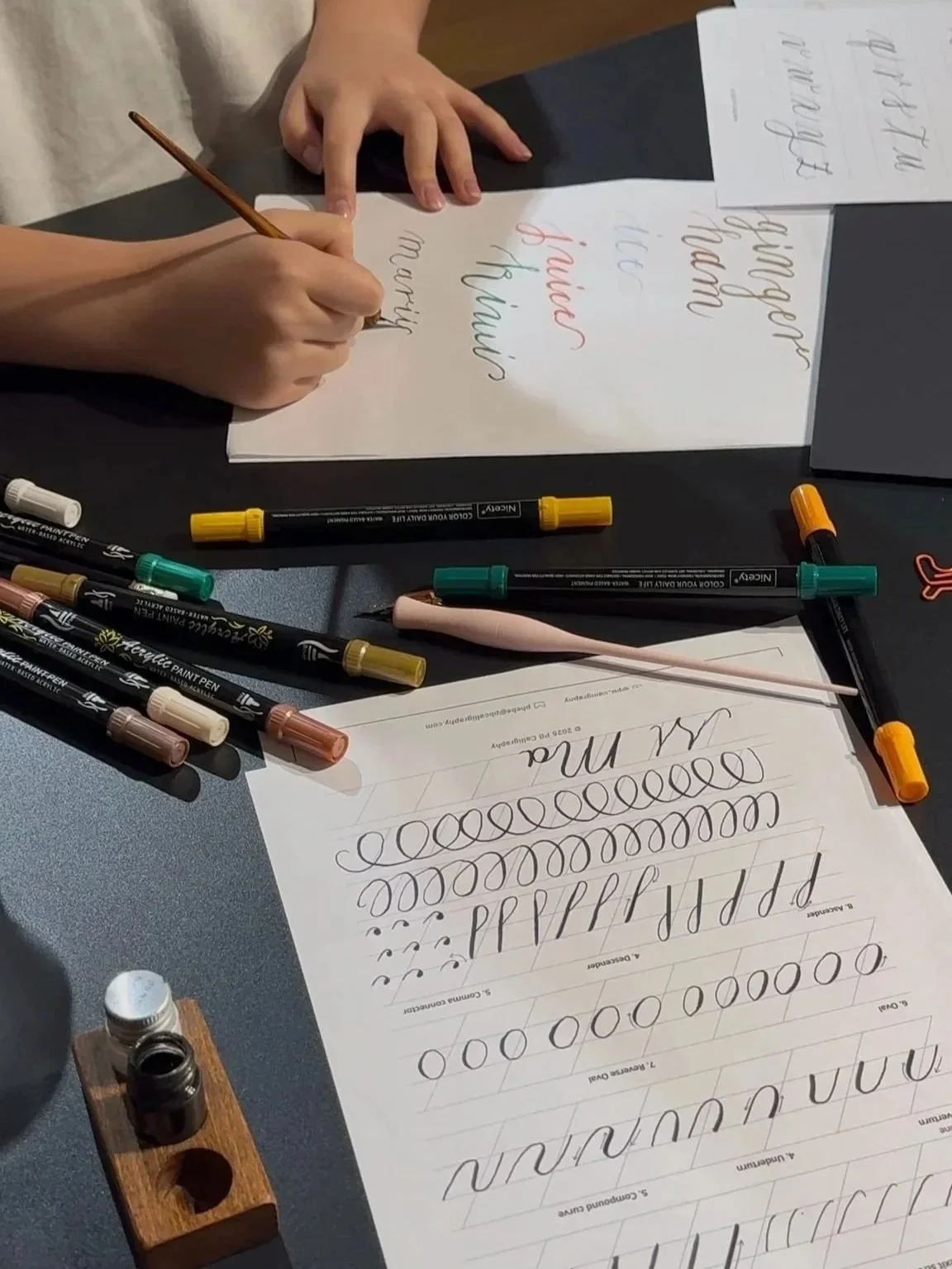 A person practicing calligraphy with various pens, a sheet of paper with cursive letters, and a child's hand resting on the table.
