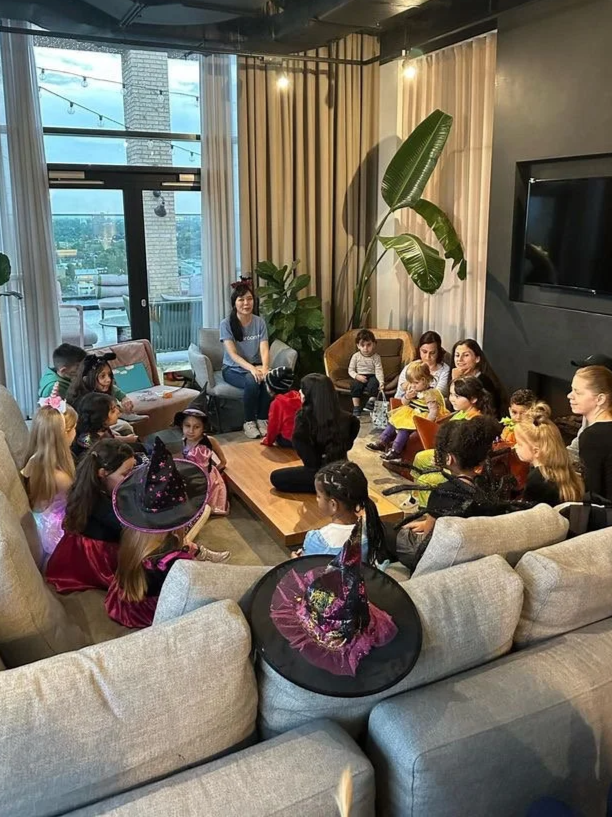 Group of children and adults gathered in a living room for a Halloween party, with some kids wearing witch hats, and a black witch hat with a pink ribbon on a gray sofa.