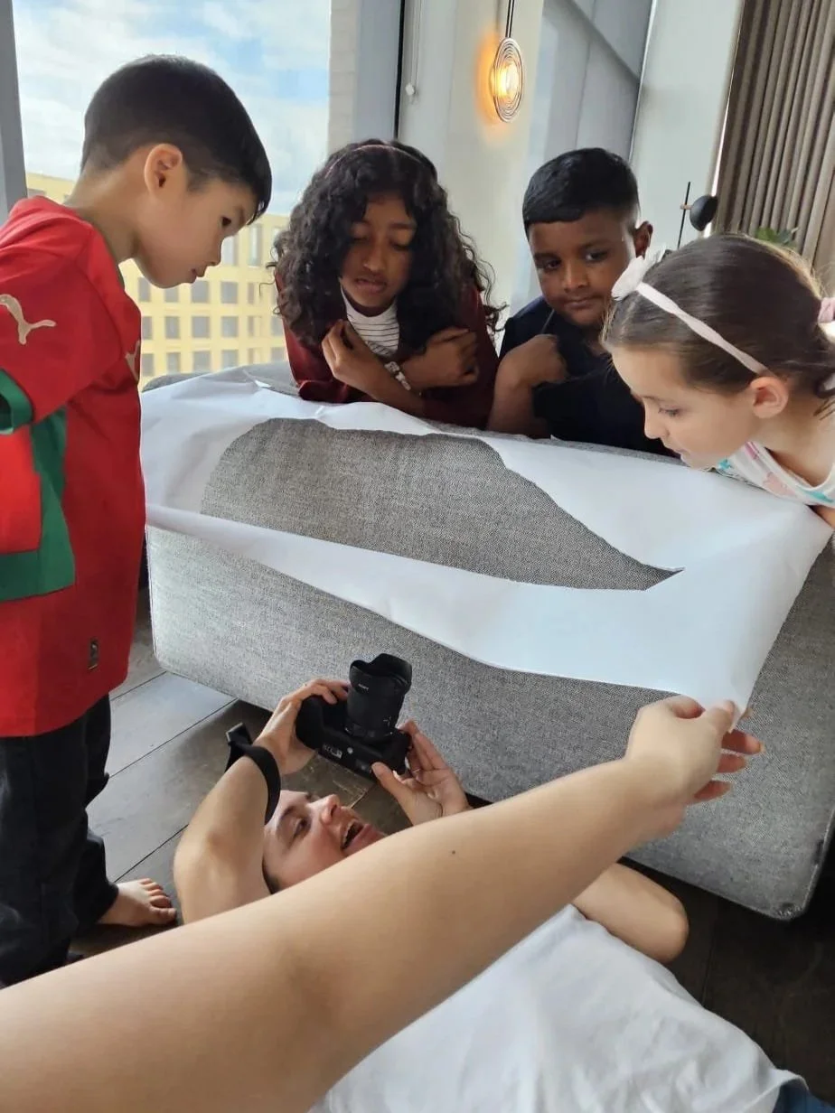 A group of children looking at a large printed image on a bed, while an adult lies underneath, taking a photo of the scene with a camera.
