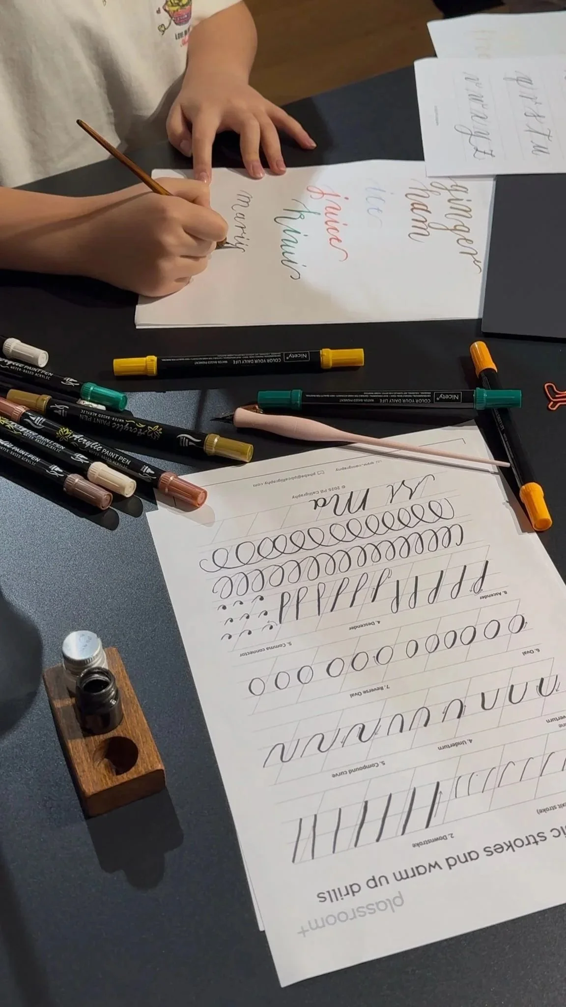 Child practicing calligraphy with various pens, writing on practice sheets with cursive letters.