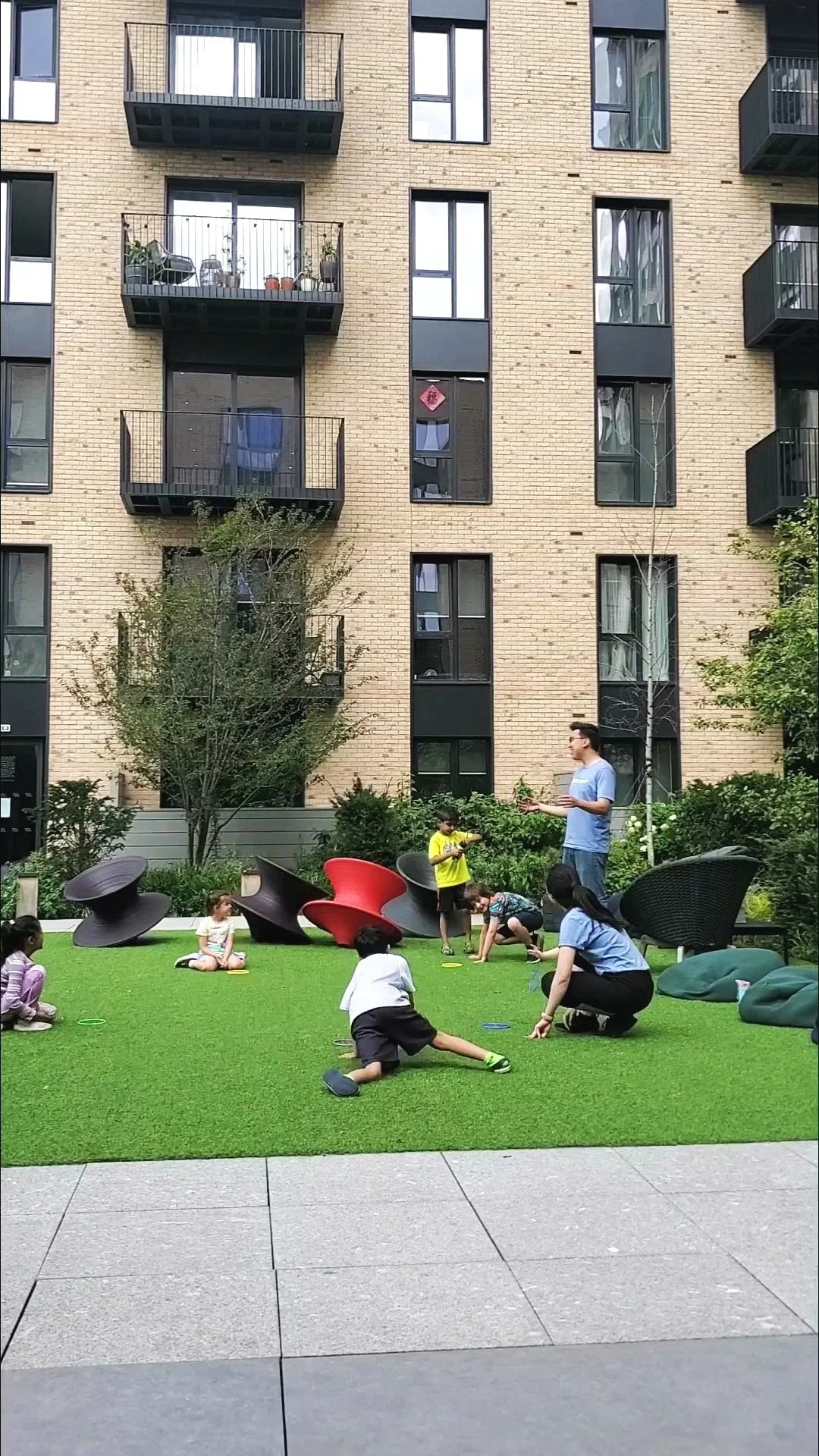 Children and adults playing and engaging in activities on a grassy area in an urban courtyard, surrounded by modern brick apartment buildings with balconies.