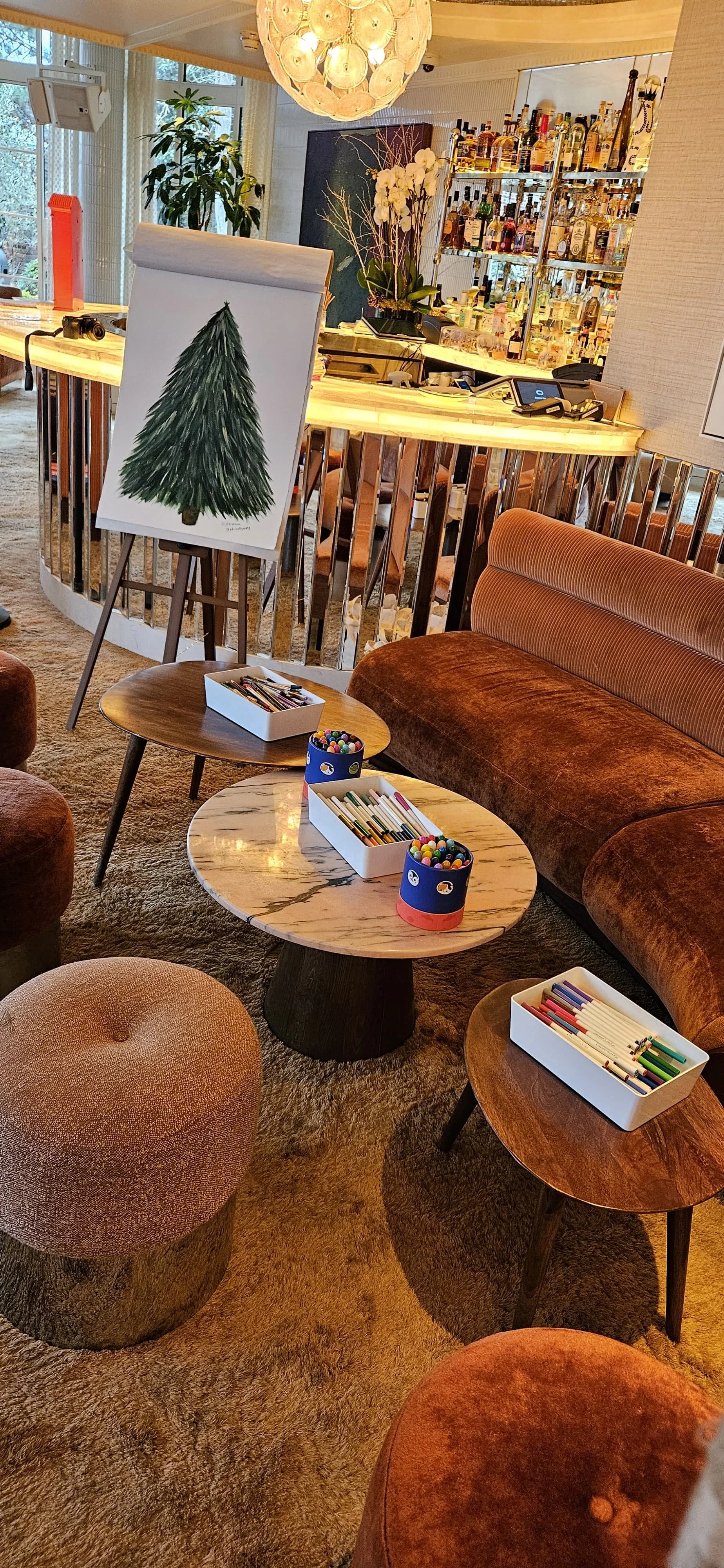 Interior of a cozy lounge area with tables, a velvet sofa, colorful chairs, and art supplies. A painting of a Christmas tree on an easel is at the center, with a bar in the background and a large decorative light fixture overhead.