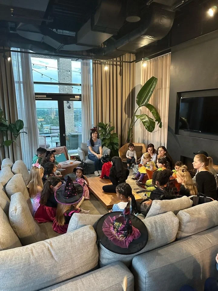 Children gathered in a cozy indoor space around a low table, with a woman leading an activity. Some children are dressed in costumes, including witches' hats. The room has large windows, potted plants, and a television mounted on the wall.