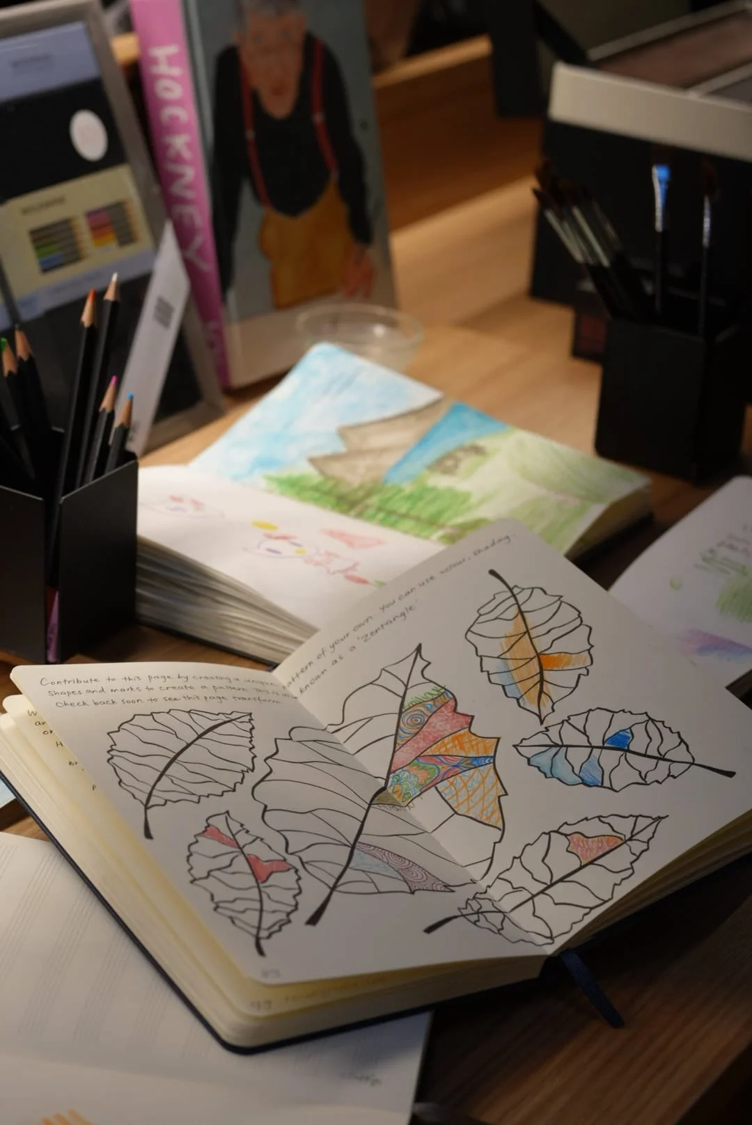 Open sketchbook with line drawing of leaves, some colored with patterns. Background shows painted landscape with mountains, grass, and sky.