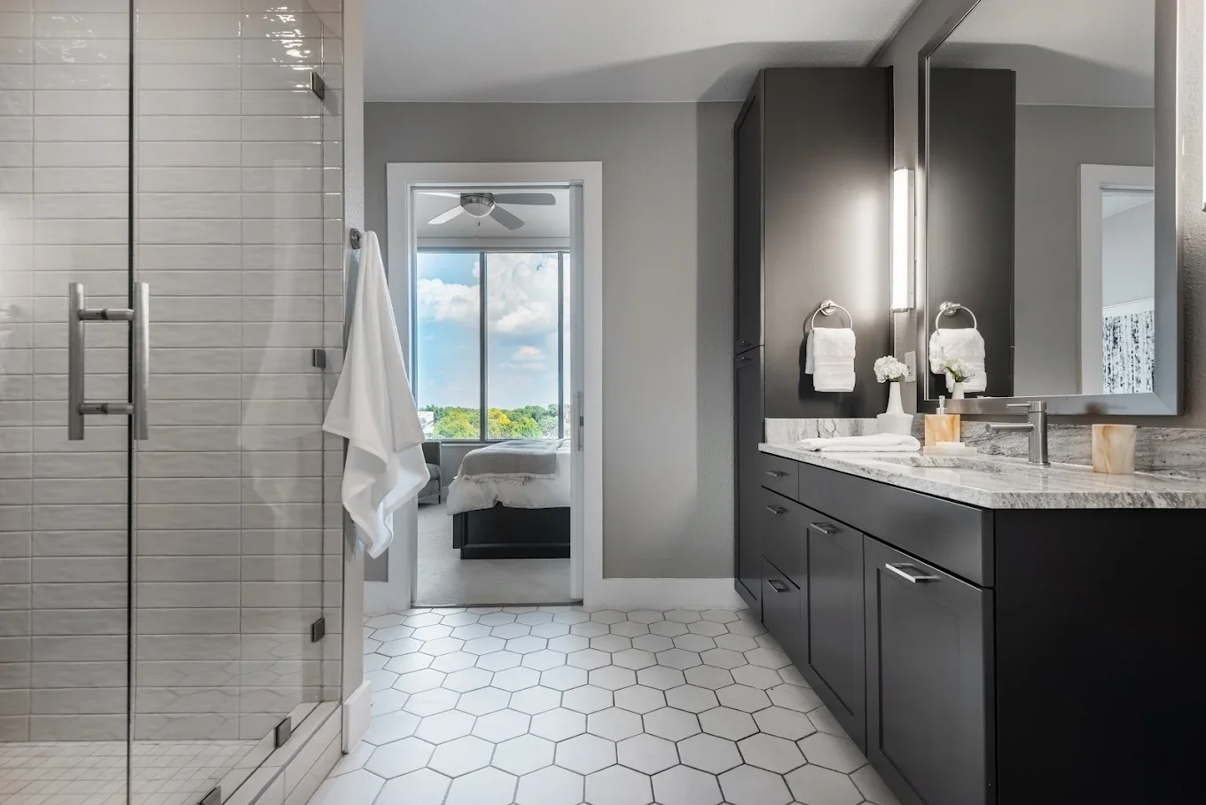 Modern bathroom with a glass shower door, dark vanity with a marble countertop, large mirror, and a bedroom with a view of greenery outside visible through a doorway.