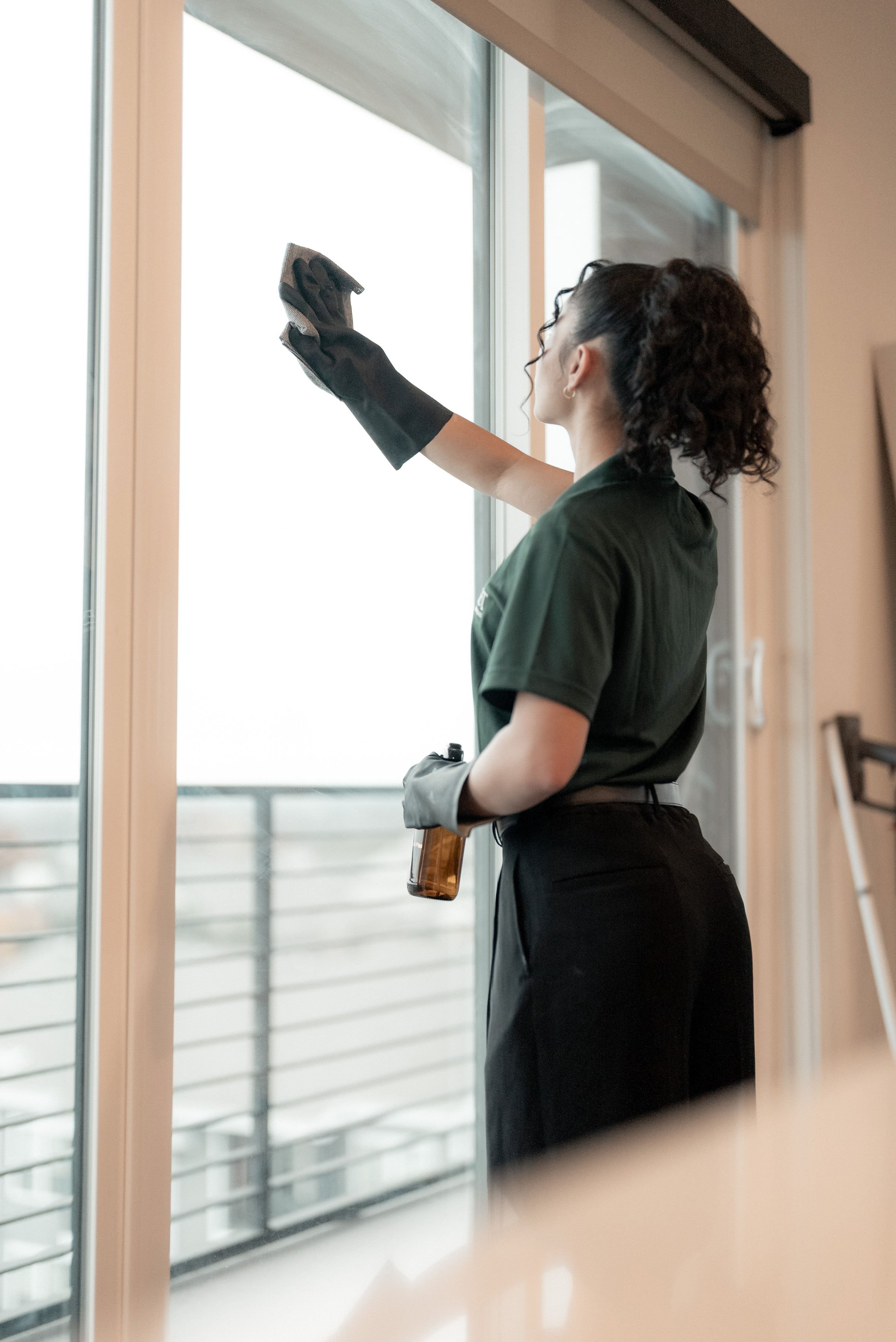A woman cleaning a glass sliding door with a cloth, wearing black gloves and holding a spray bottle in her other hand.