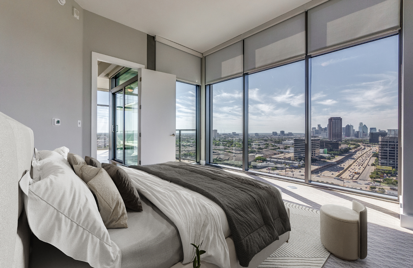 Modern bedroom with large floor-to-ceiling windows showing a city skyline and highway, bed with white and gray bedding, and a small round white ottoman near the window.