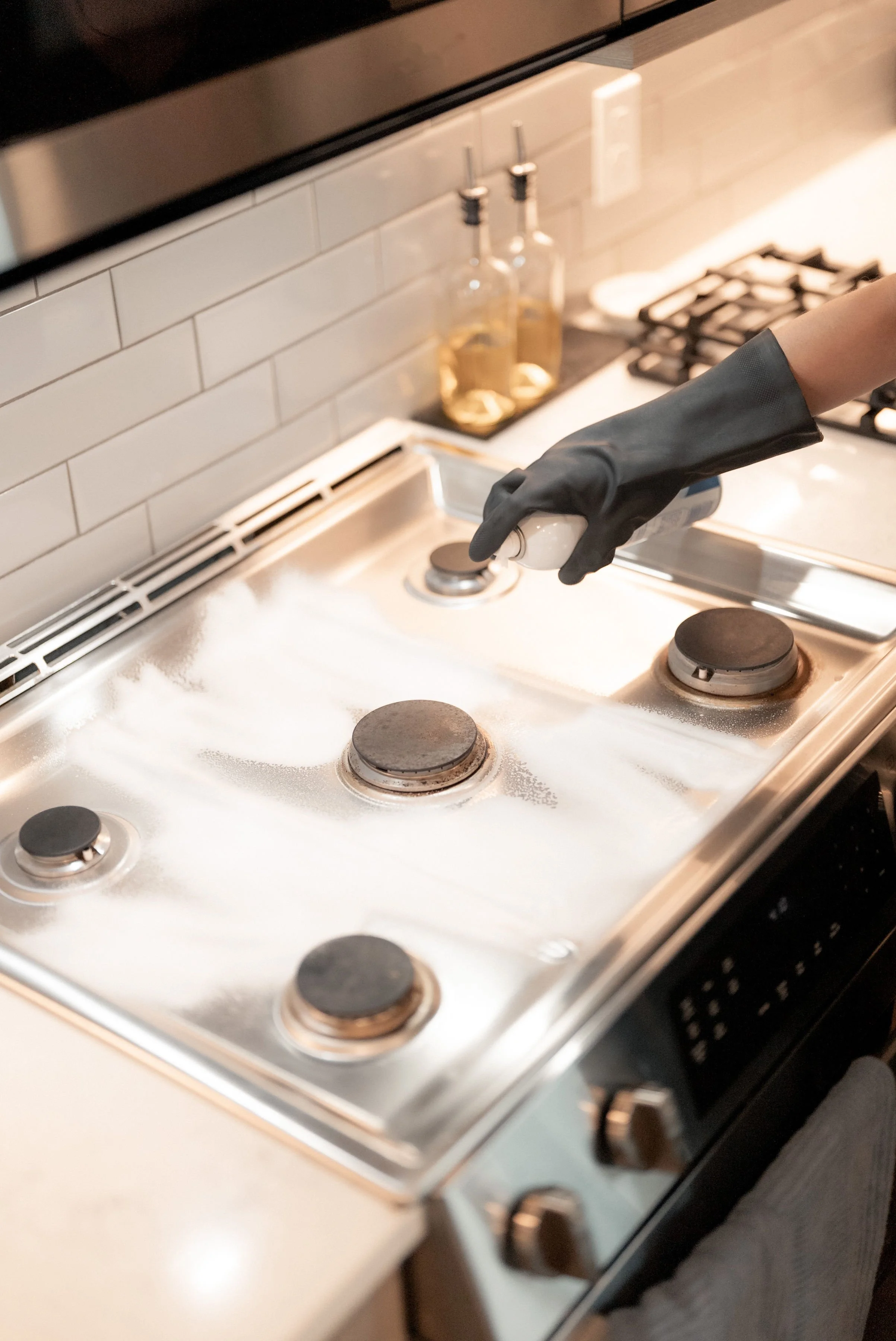 Hand wearing a black glove spraying cleaning solution onto a dirty stovetop with burnt-on residue.