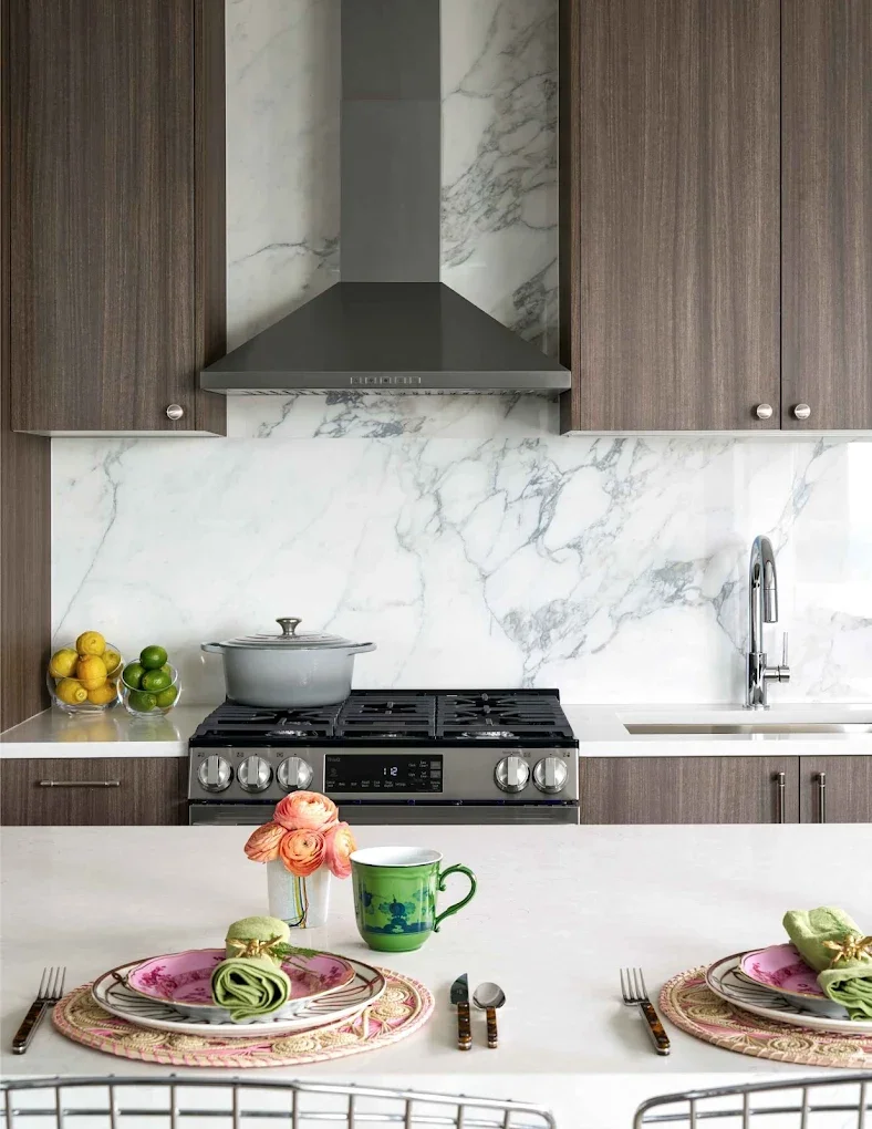 Kitchen with marble backsplash, dark wood cabinets, stainless steel gas stove, pot on stove, bowl of lemons and limes on countertop, sink with faucet, dining table with pink and green dishware, pink roses in a vase, green coffee mug, utensils, and placemats.