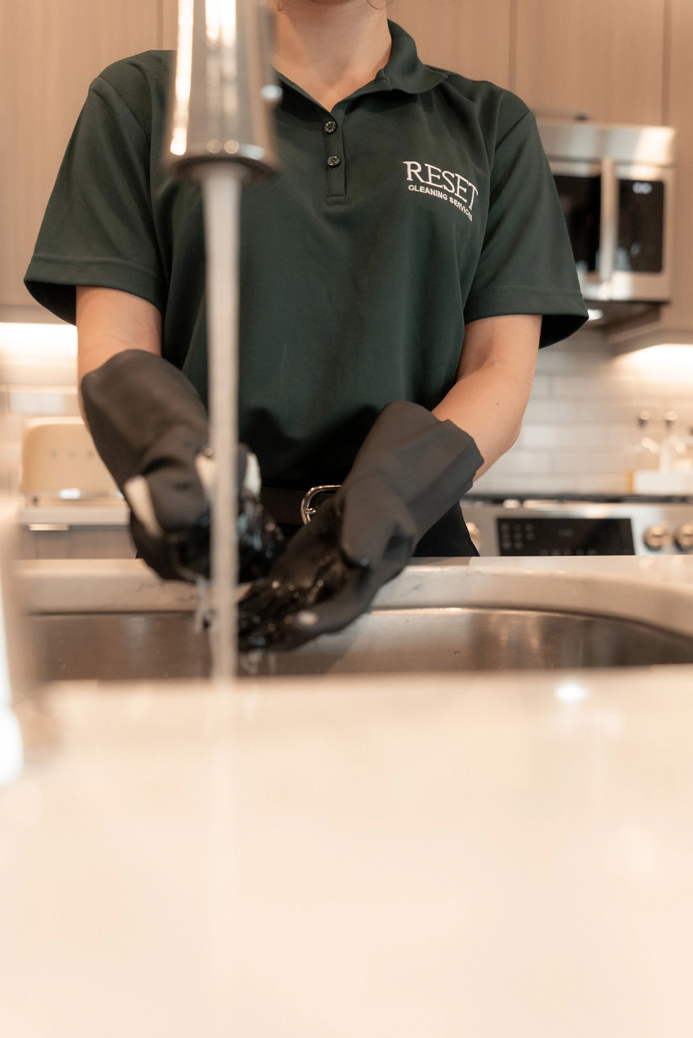 Person washing dishes wearing black rubber gloves and a green polo shirt with 'RESET' logo in a kitchen.