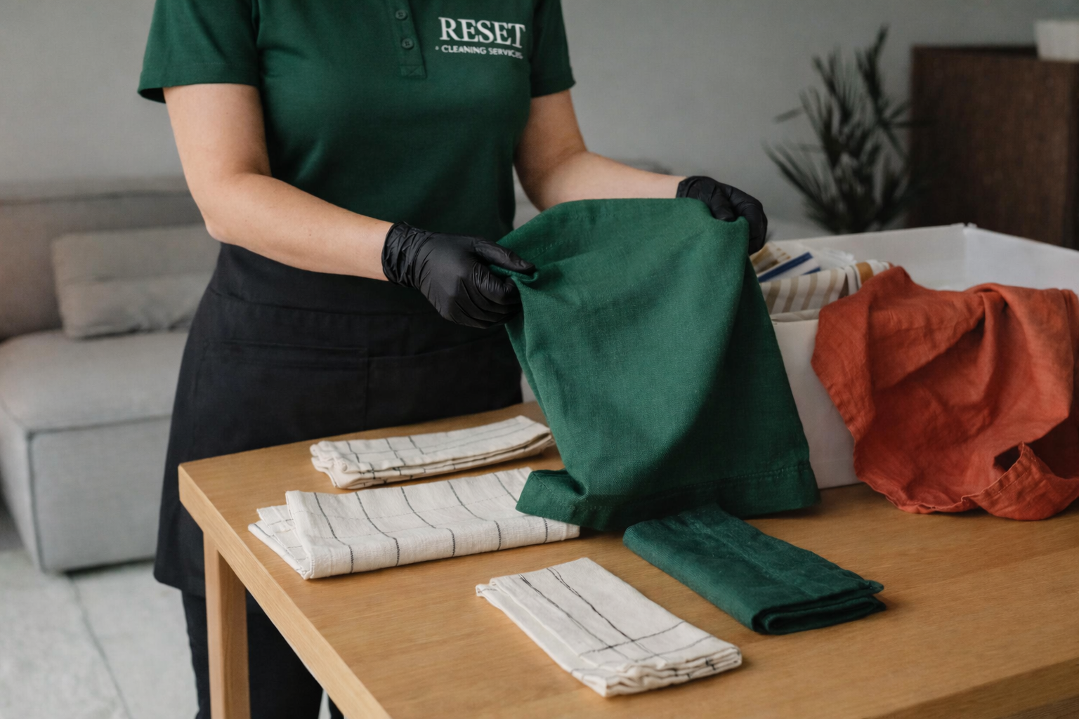 Person in green shirt and black gloves folding green cloth on a wooden table with folded towels and fabric pieces.