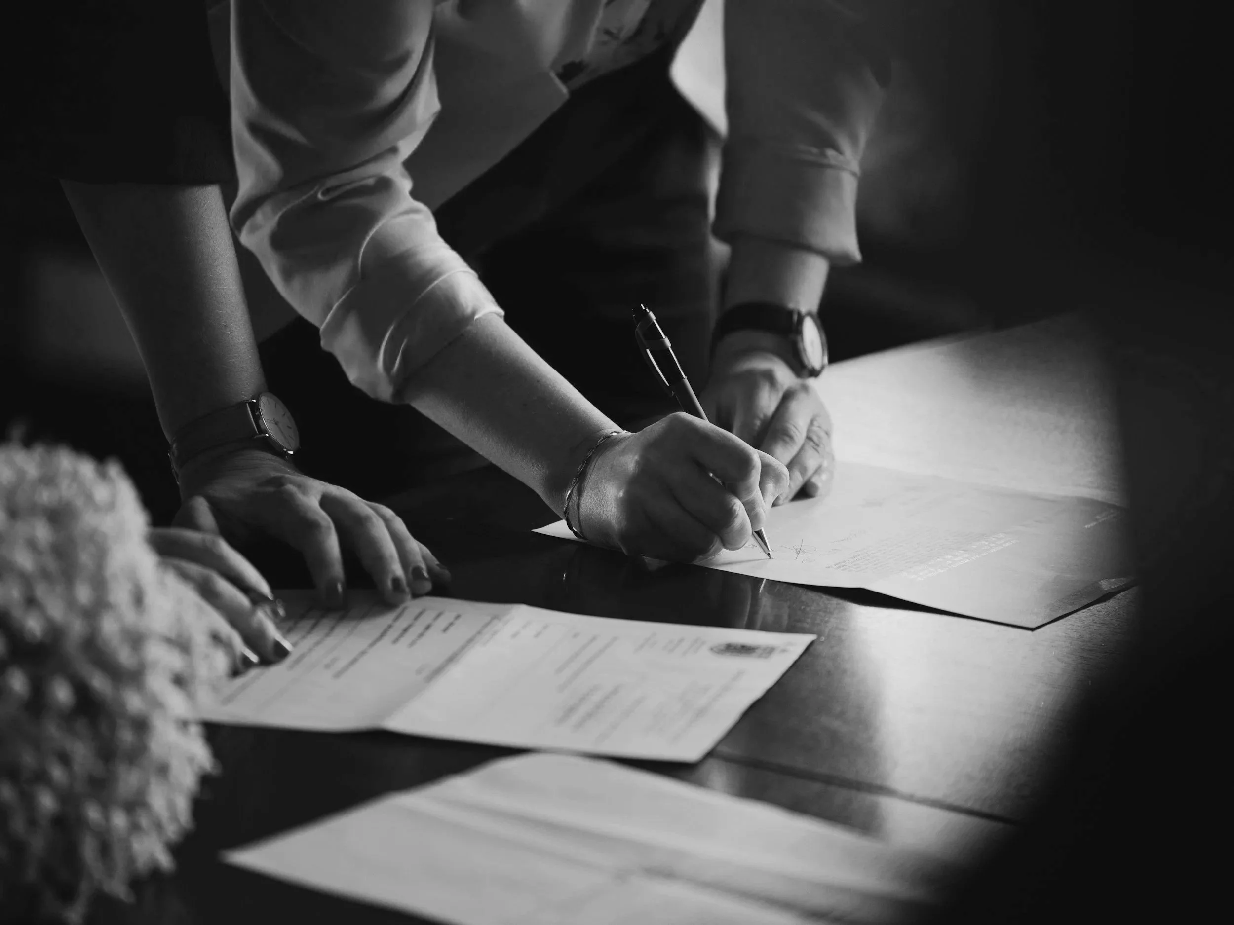 A person writing on paper at a legal table, with documents and a bouquet of flowers nearby, black and white photo.