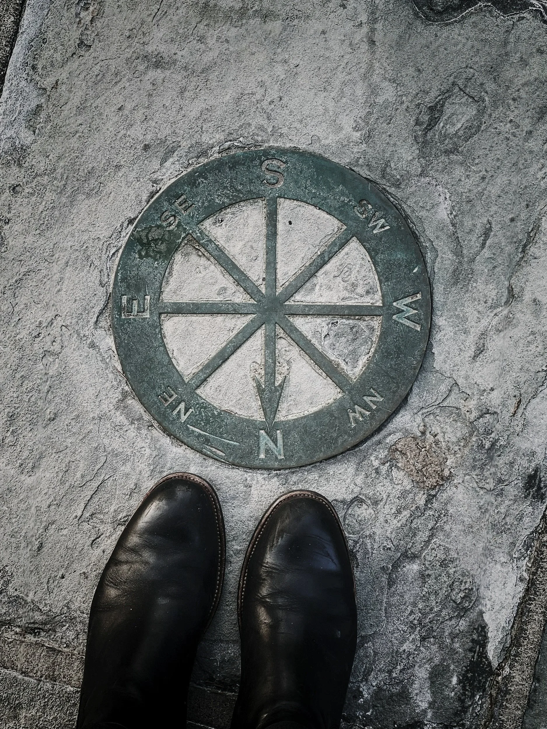 A person wearing black shoes standing on a stone floor, looking down at a circular metal compass embedded in the ground, showing the cardinal directions.