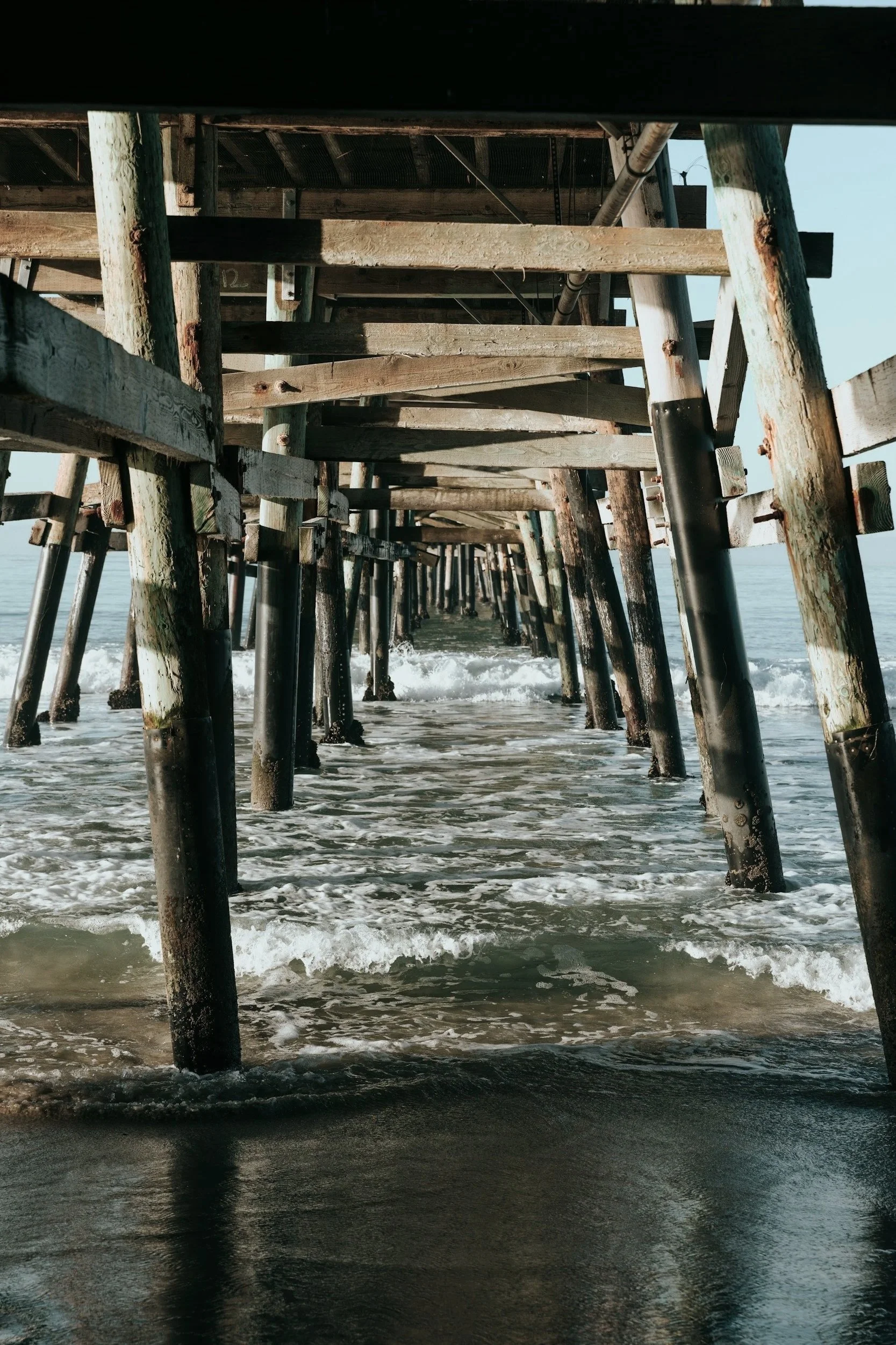View underneath a wooden pier extending over the ocean with waves crashing against the pilings.