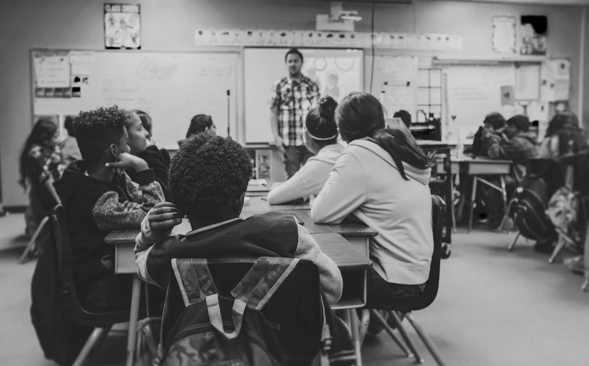 A classroom with students sitting at desks facing a teacher at the front, who is standing near a whiteboard. The students are attentive, and the classroom has educational posters and supplies.