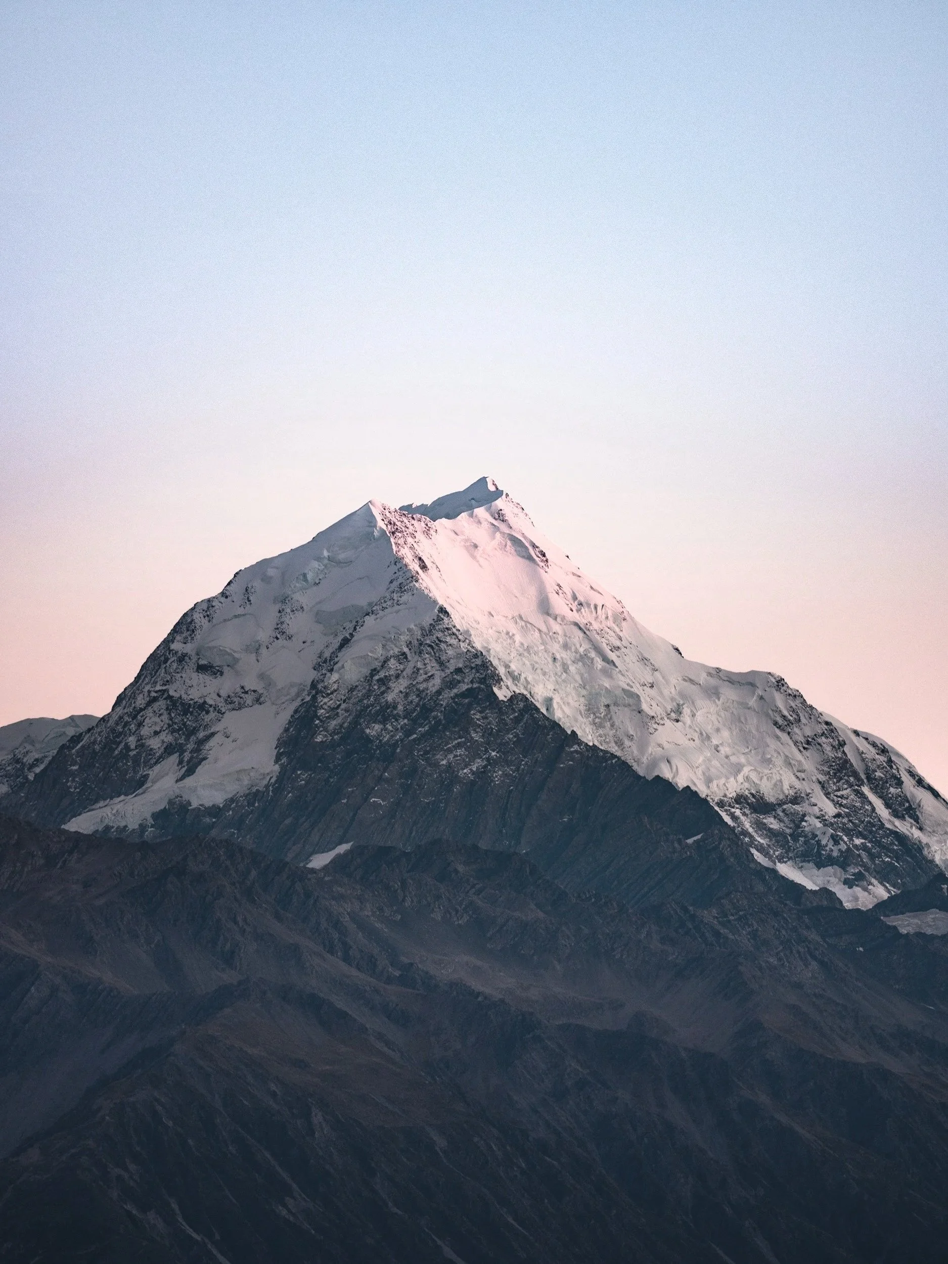 Snow-capped mountain peak with rugged dark lower slopes under a pale sky.