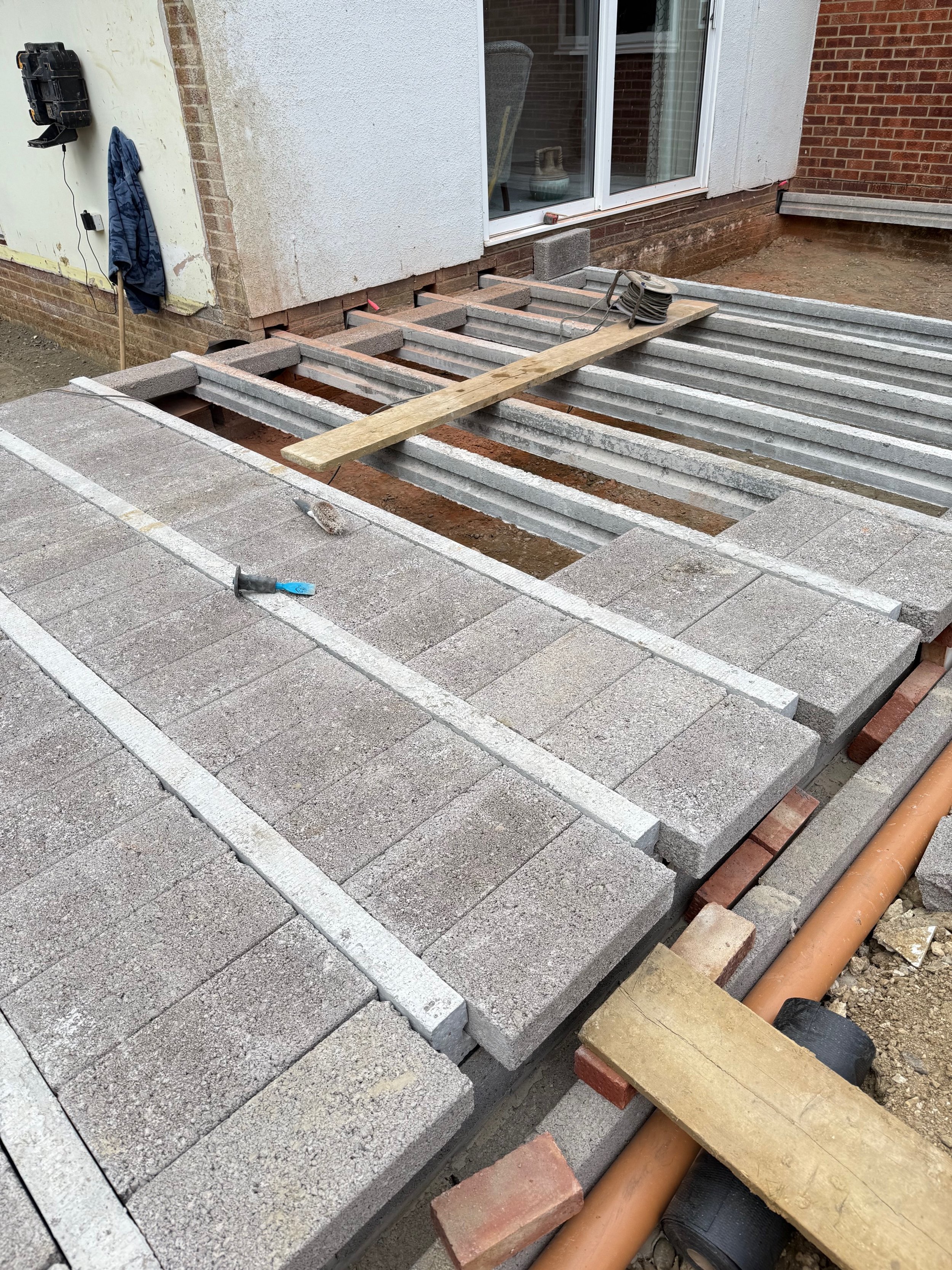Construction site with paving stones and concrete slabs being laid, with tools and wooden support beams present, next to a house with a sliding glass door.