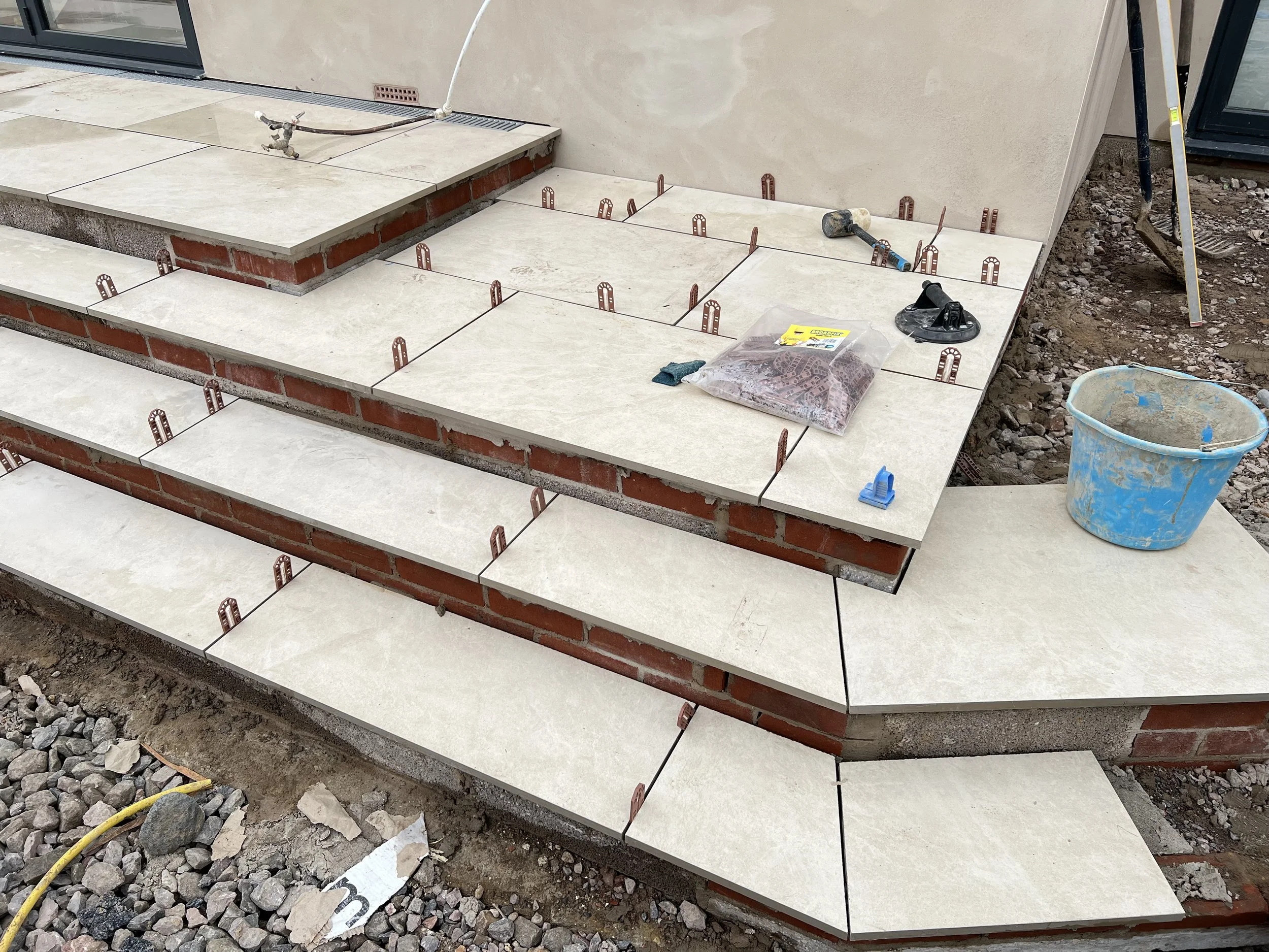 Construction site with beige tile steps partially installed, tools, a bucket of construction material, and exposed brick and dirt.