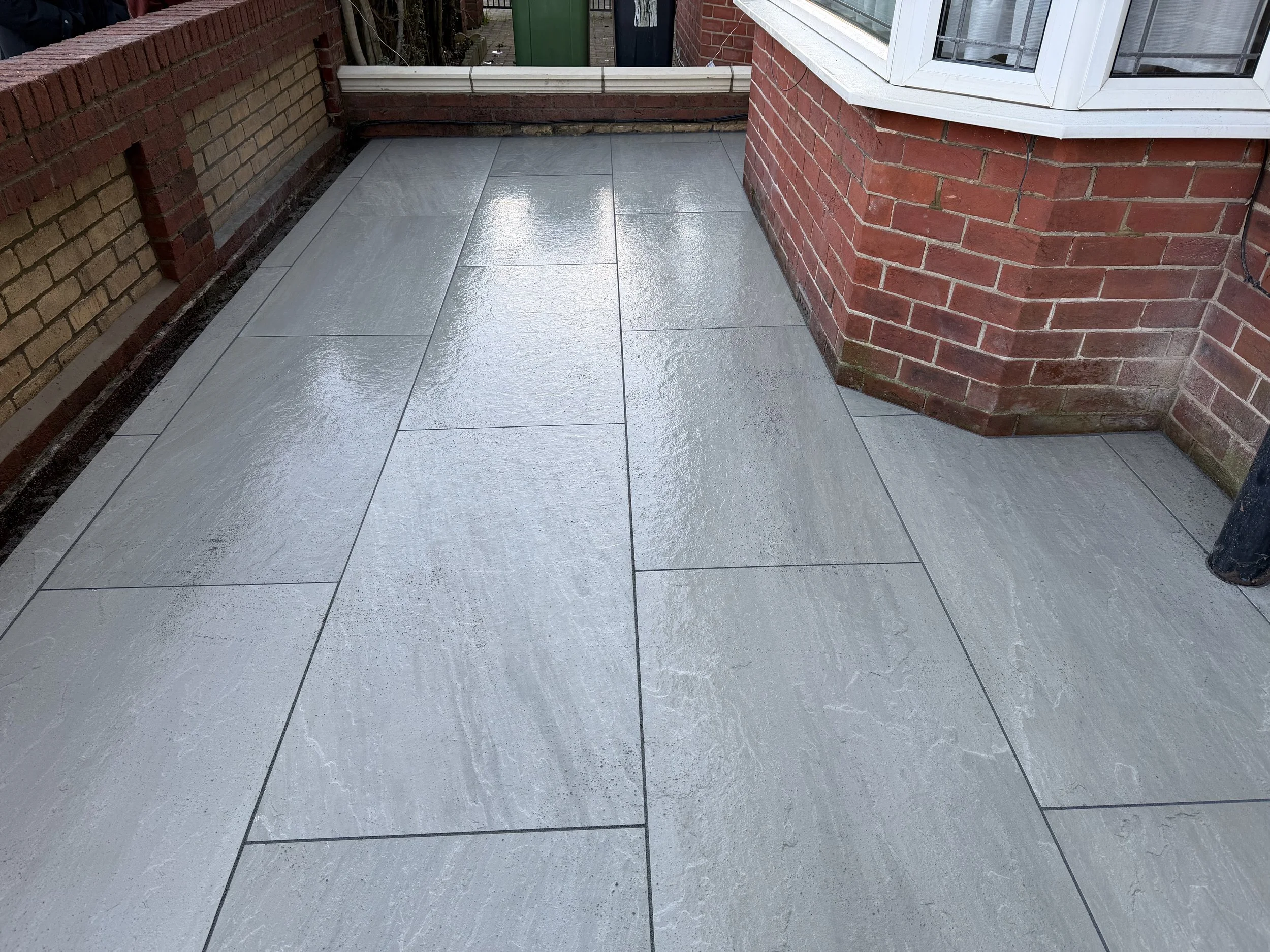 Freshly laid light gray paving stones on a balcony or patio area, bordered by red brick walls and a small section of a bay window.