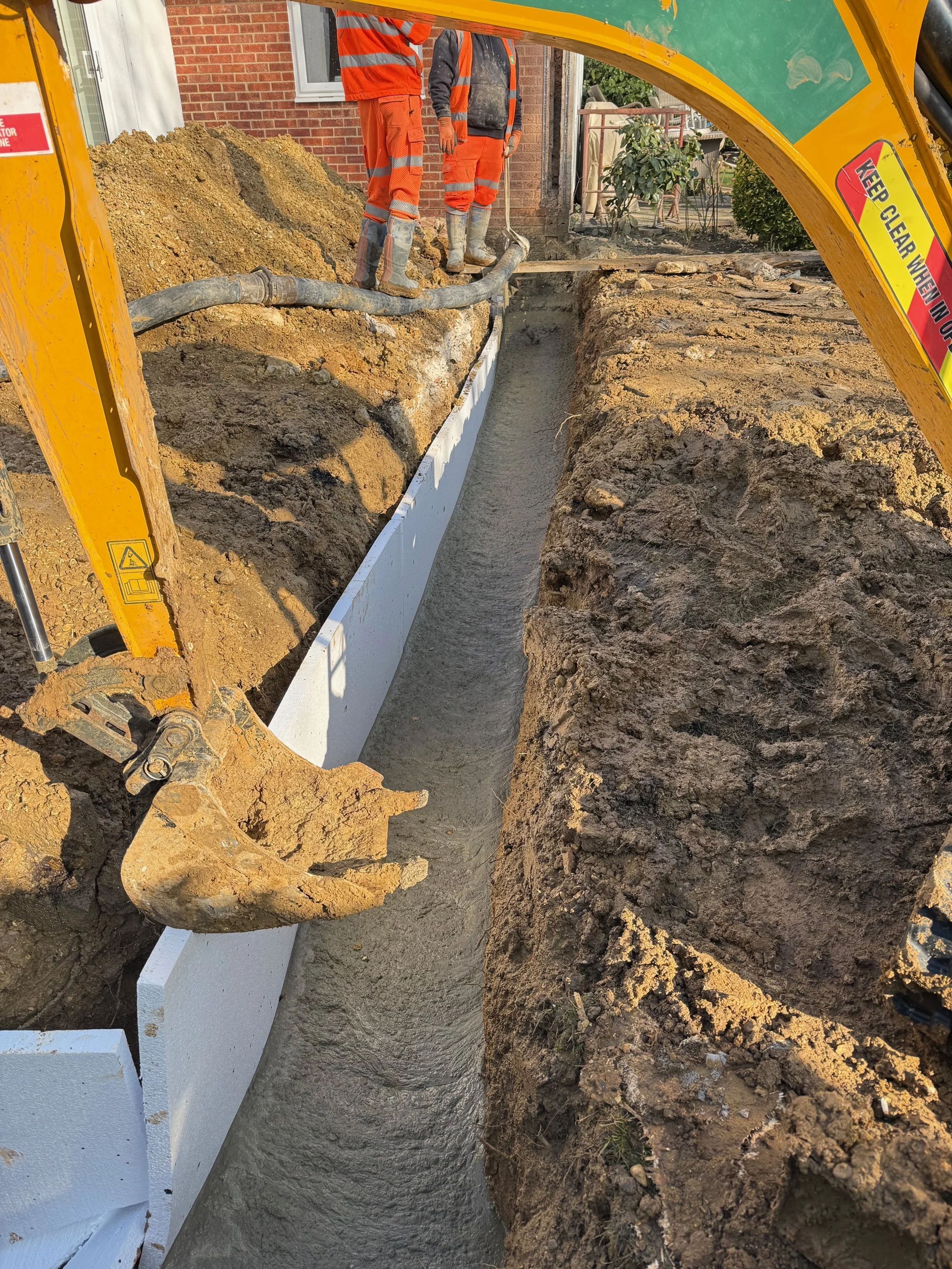 Construction workers in orange safety gear standing in a trench, pouring concrete with a yellow excavator nearby, in a residential area.