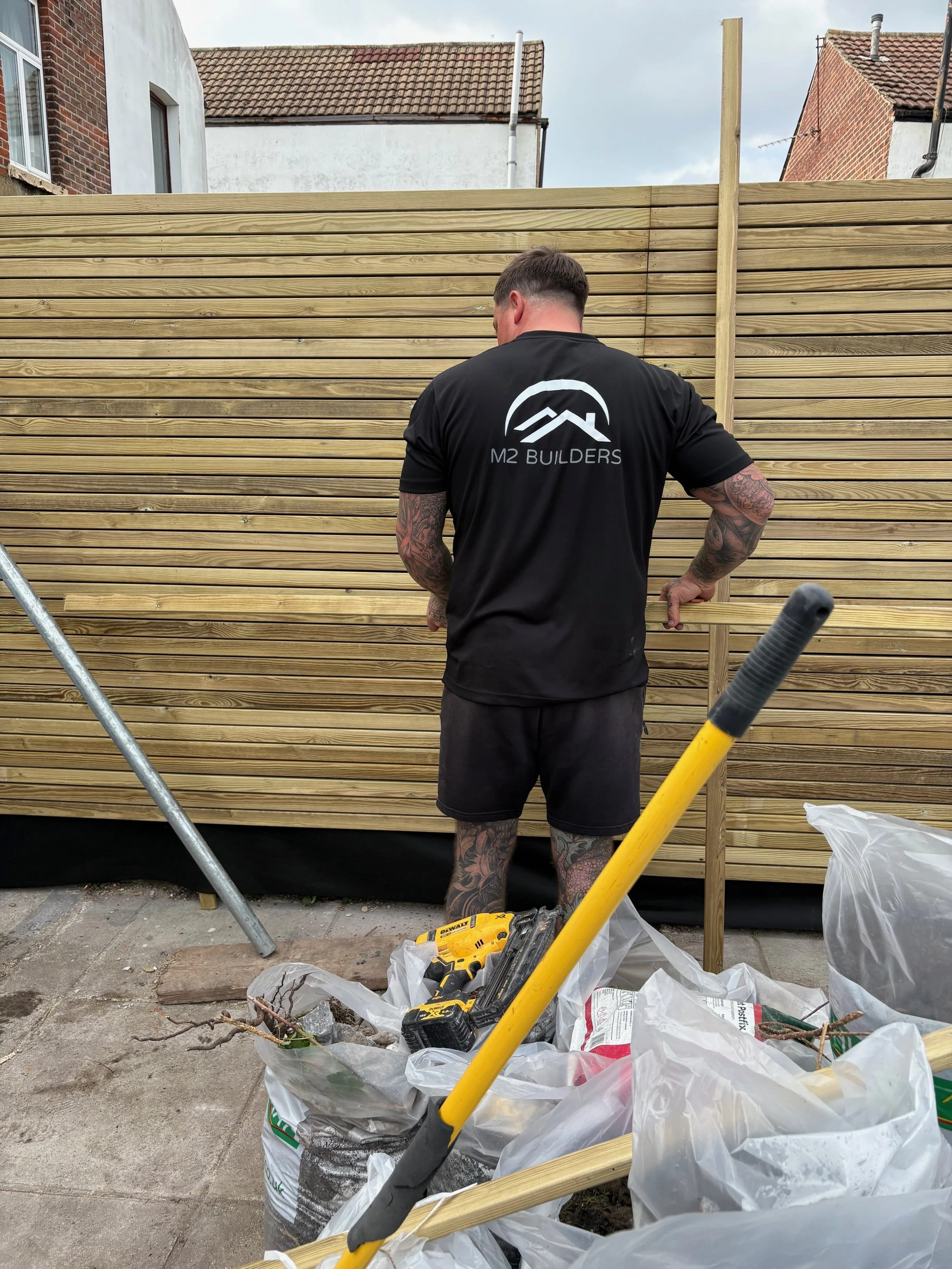 A man with tattoos on his arms and legs is working on building a wooden fence in a backyard. He is wearing a black T-shirt with the logo and name 'M2 Builders' on the back. There are construction tools and materials, including a yellow cordless drill