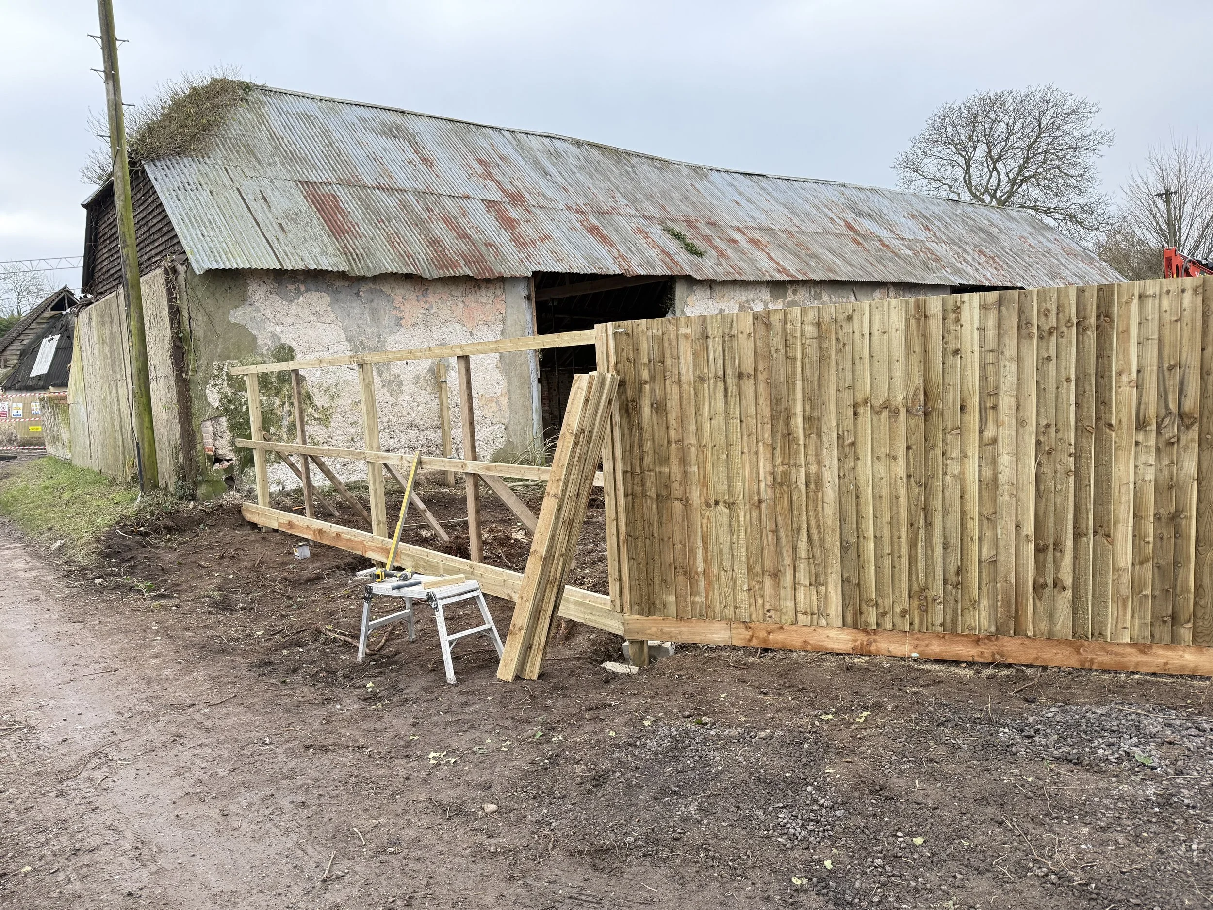 A rural construction site where a new wooden fence is being built in front of an old, weathered barn with a metal roof. Construction tools sit on a small ladder, and the ground is muddy.