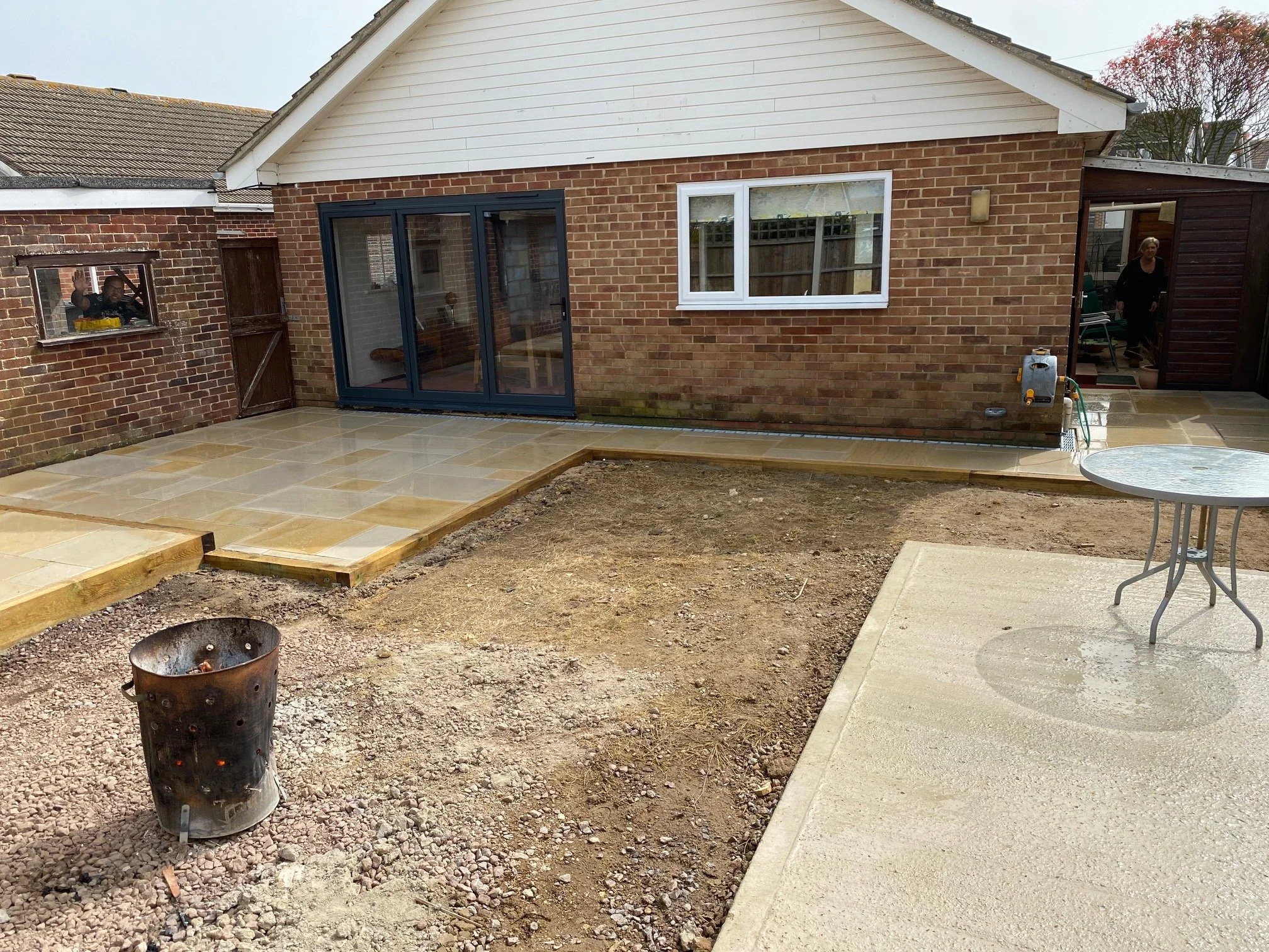 Backyard with newly laid stone patio and an unfinished dirt area prepared for additional paving or turf, with a round glass table on the paving. There is a brick house with white trim and a sliding glass door leading inside.