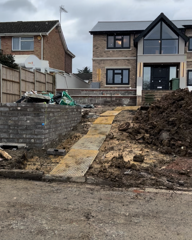 Construction site with a yellow metal ramp leading up to a modern house, muddy ground, construction debris on the left, and neighboring houses in the background.