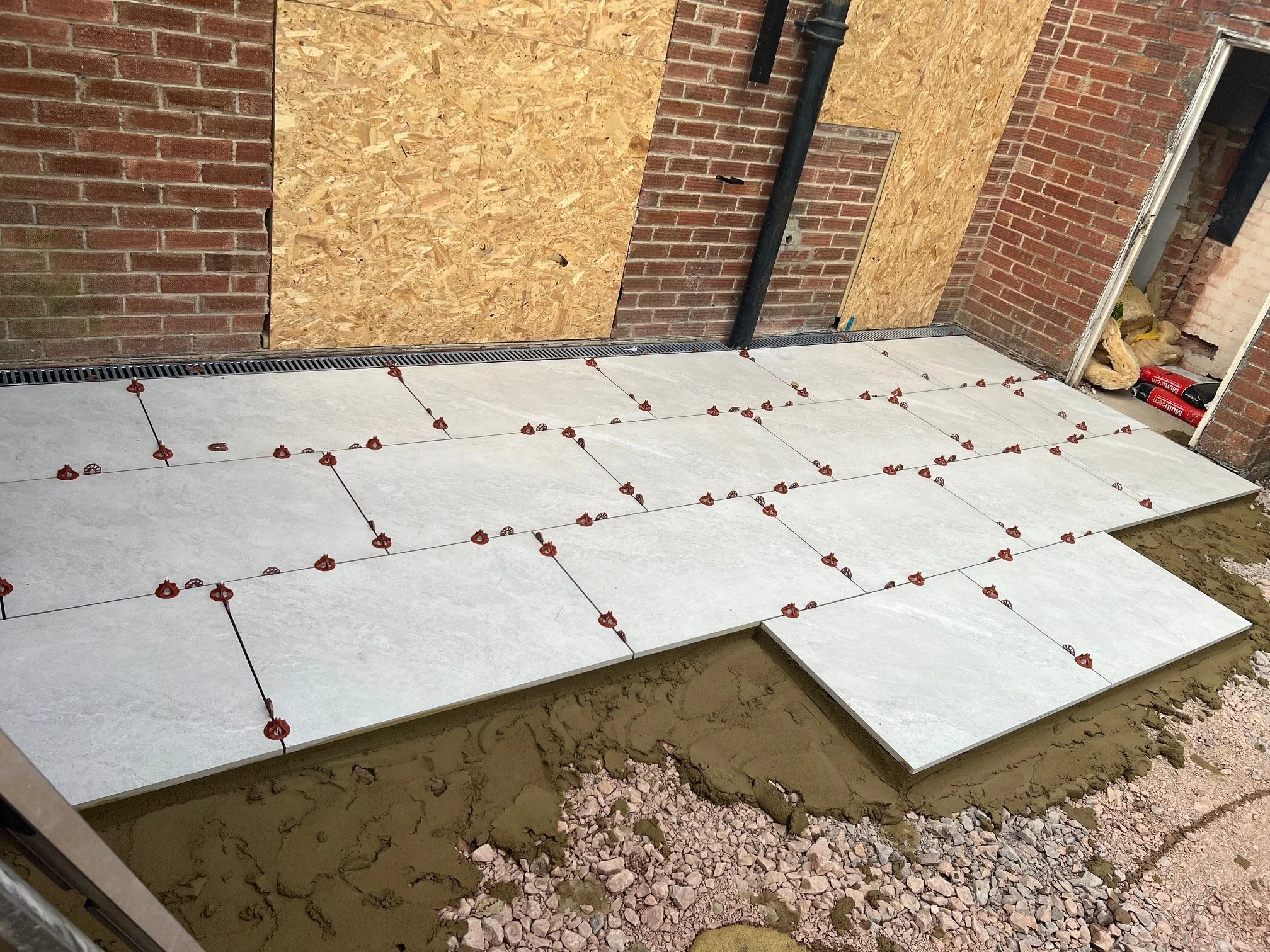 Construction site showing tiles being installed on a concrete floor outside a building with brick walls. Some tiles are already placed, and others are being aligned with spacers.