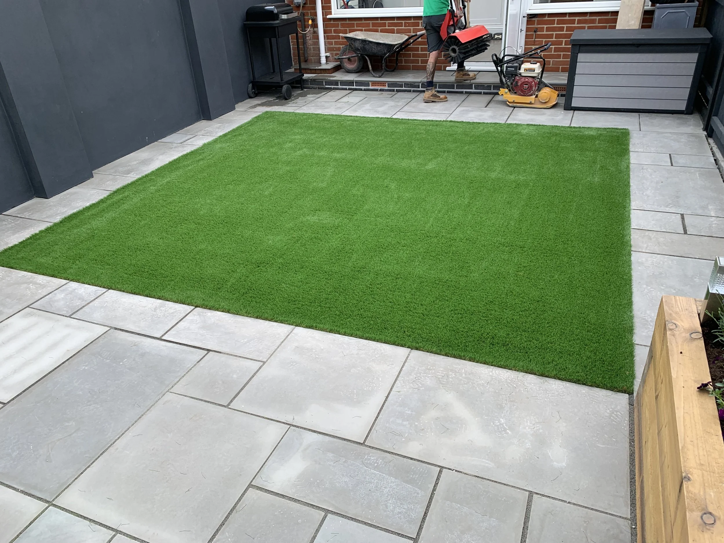 Green artificial grass rug on a stone patio, with some gardening and construction tools in the background.