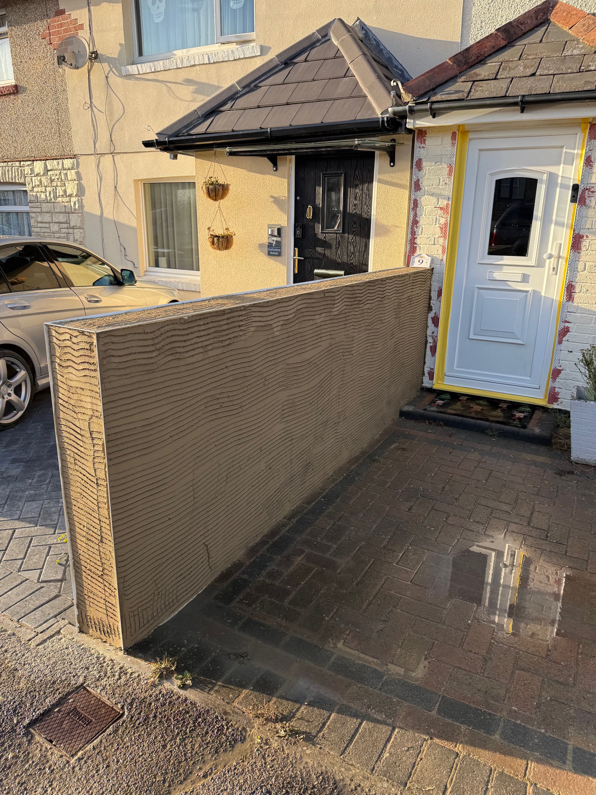 Front entrance of a house with a black door and a white door, a brick and concrete wall, a parked car, hanging flower baskets, and a brick patio with a puddle reflecting the house.