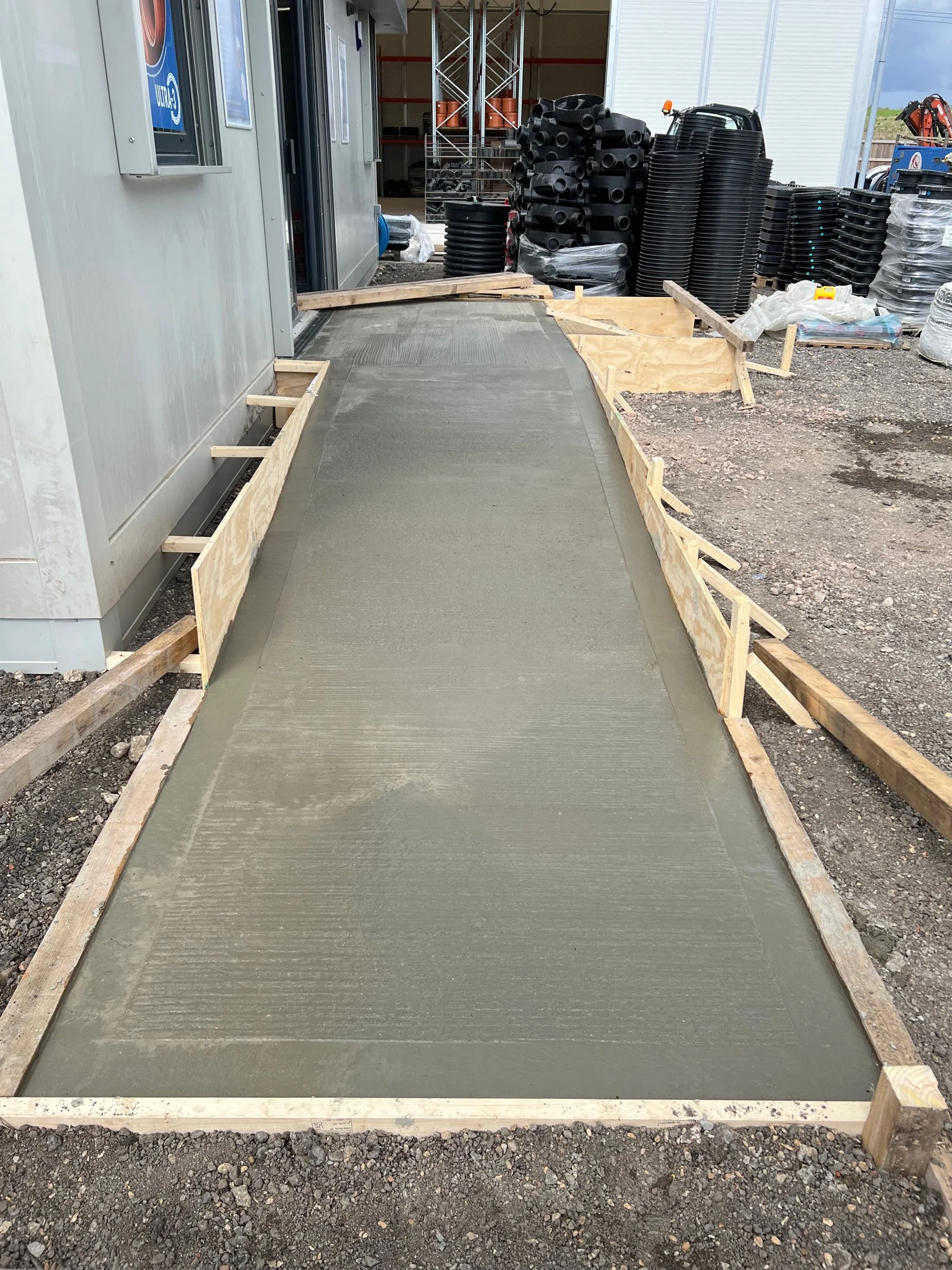 A freshly poured concrete sidewalk on a construction site, with wood form framing around the edges, adjacent to a gray building, and construction materials and equipment in the background.