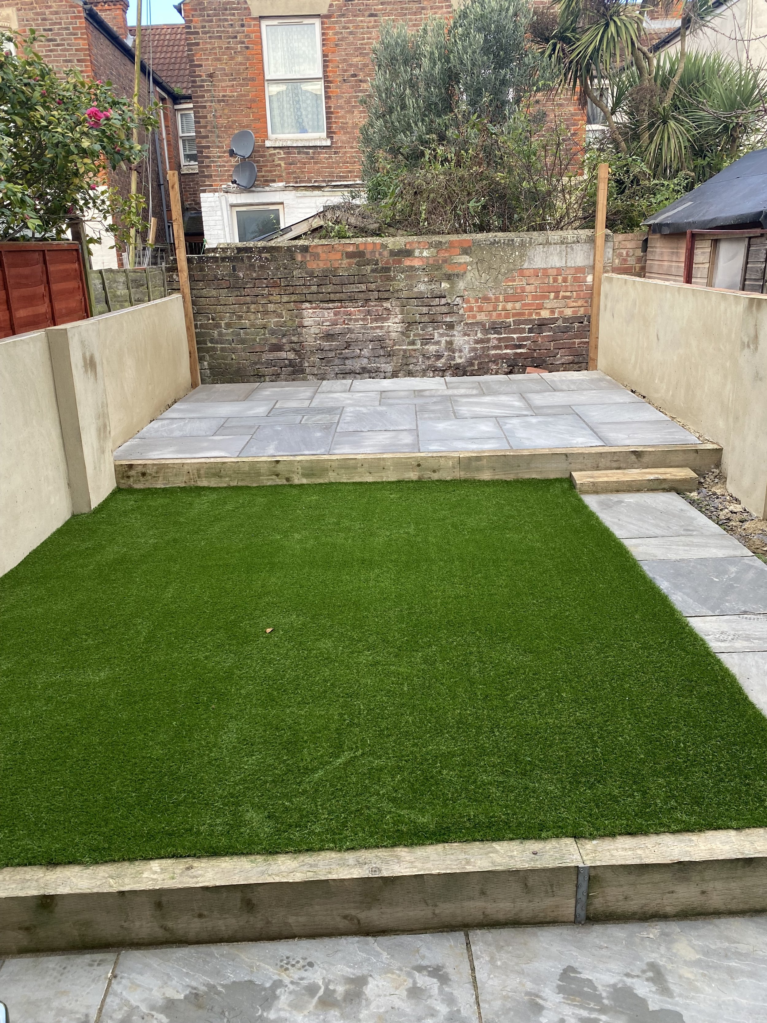 A backyard with a patch of green artificial turf, a stone patio, and a wooden border. The patio has large gray stone tiles on an elevated wooden platform, with a small set of stone steps leading down from the patio. The yard is enclosed by beige and 