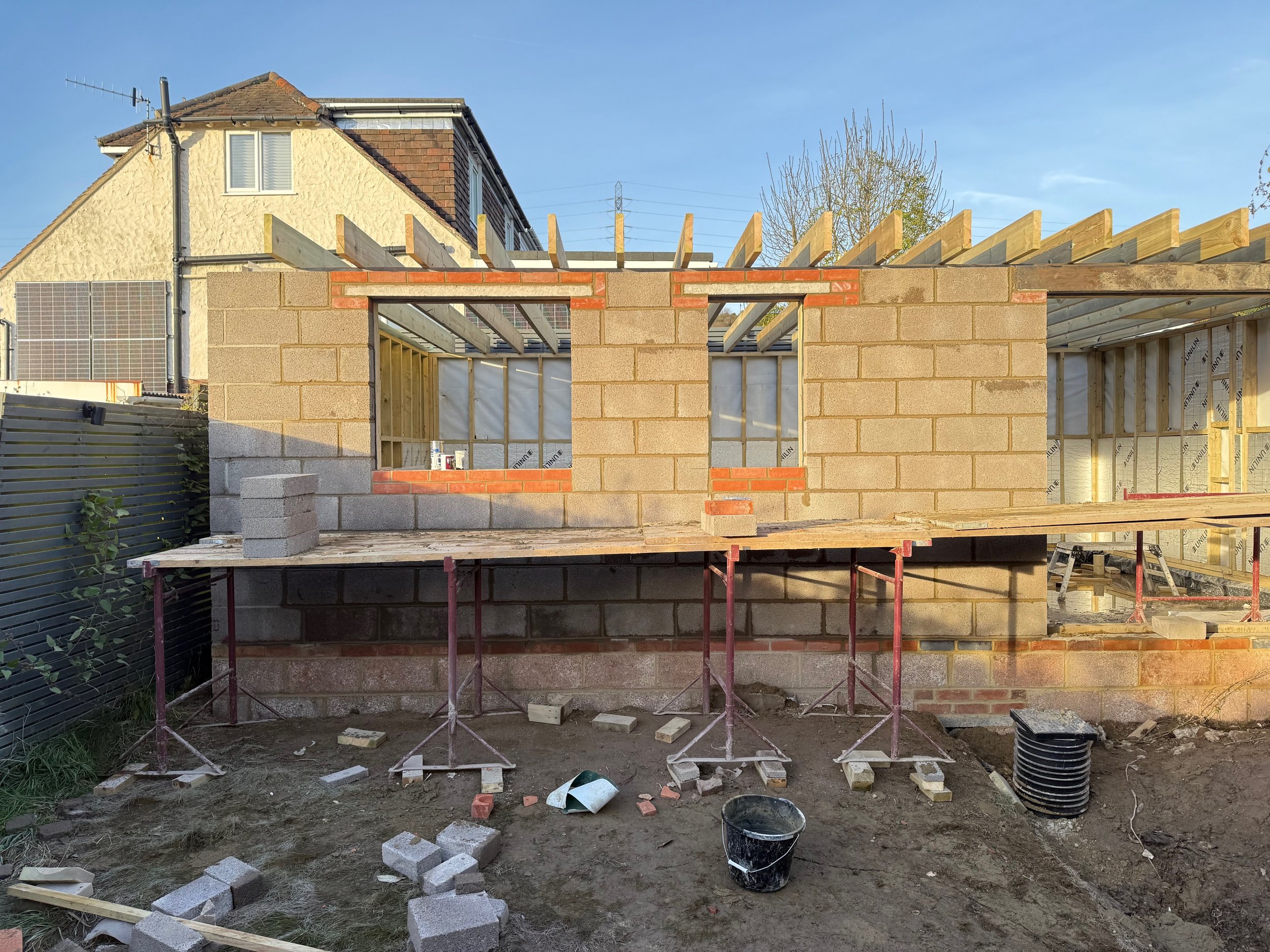 Construction site of a small building with brick and concrete walls, wooden framing at the top, window openings, and scaffolding at the ground level.