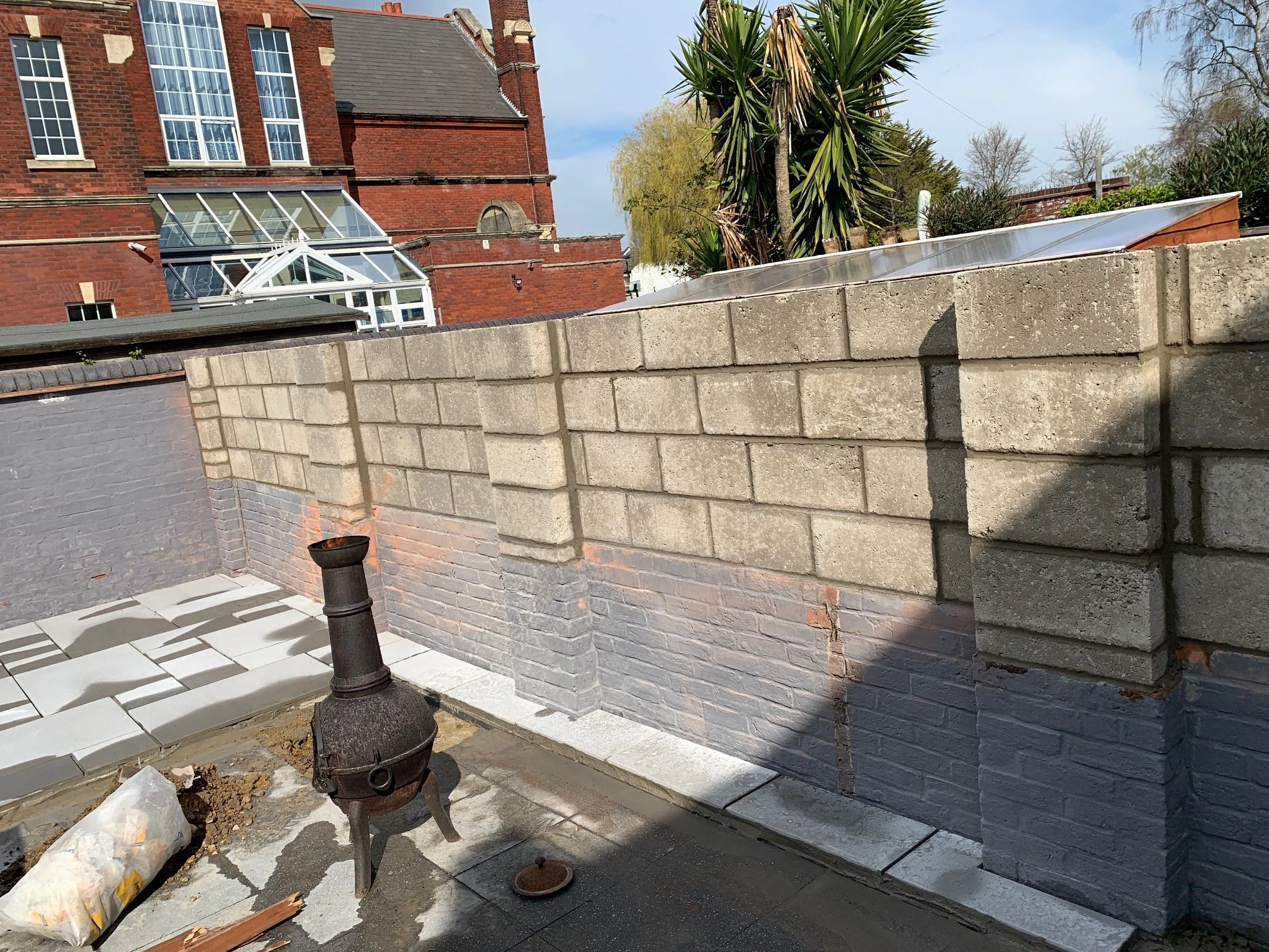View of a rooftop with a brick wall, construction materials, and a small chimney or stove, with buildings and trees in the background under a partly cloudy sky.