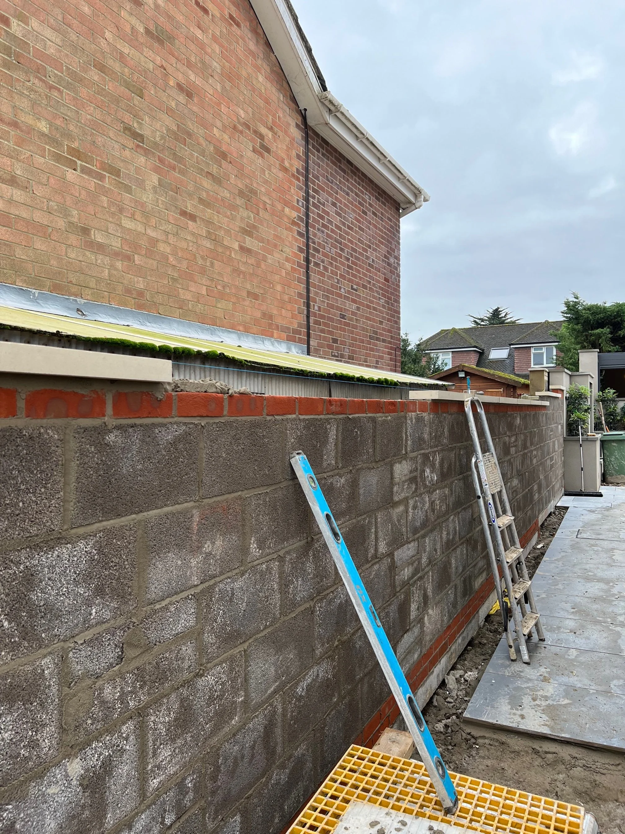 Construction site showing a concrete block wall being built, with two ladders and construction materials in the area. Part of a brick house is visible in the background under an overcast sky.