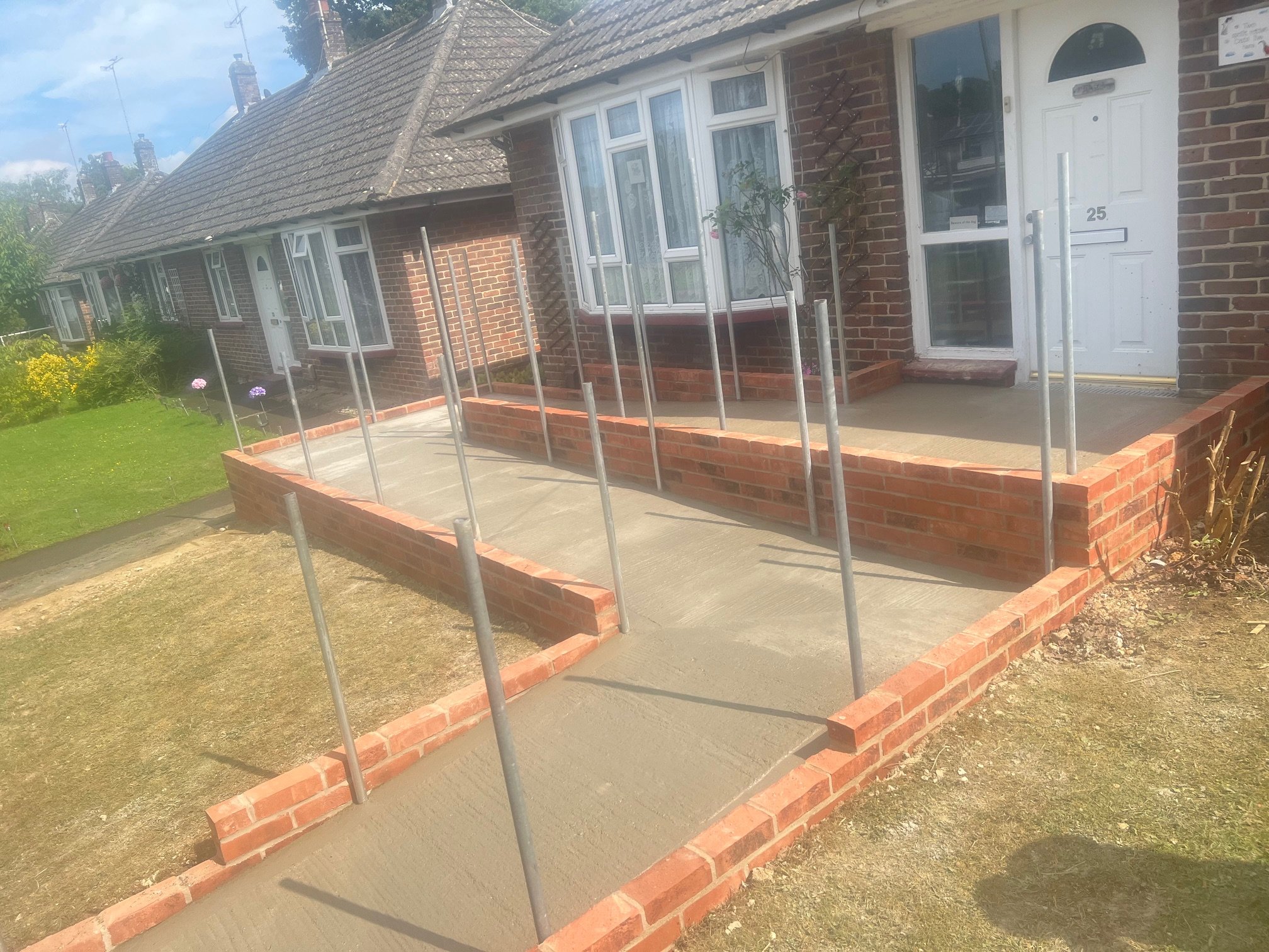 Newly constructed concrete ramp with brick border and metal handrails outside of a brick house with a white door and bay window, leading to the entrance of the house on a sunny day.