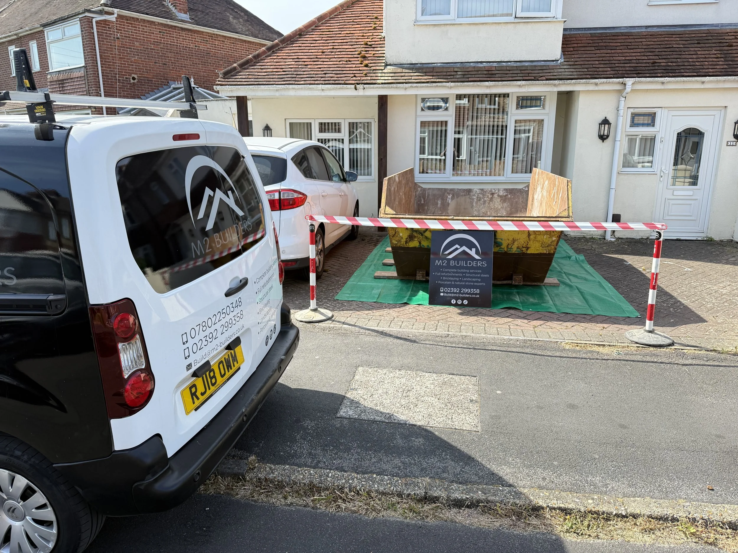Construction skip in front of a house with construction company's sign, surrounded by a barrier tape. Two parked cars, a white one and a black one, are nearby on the street.