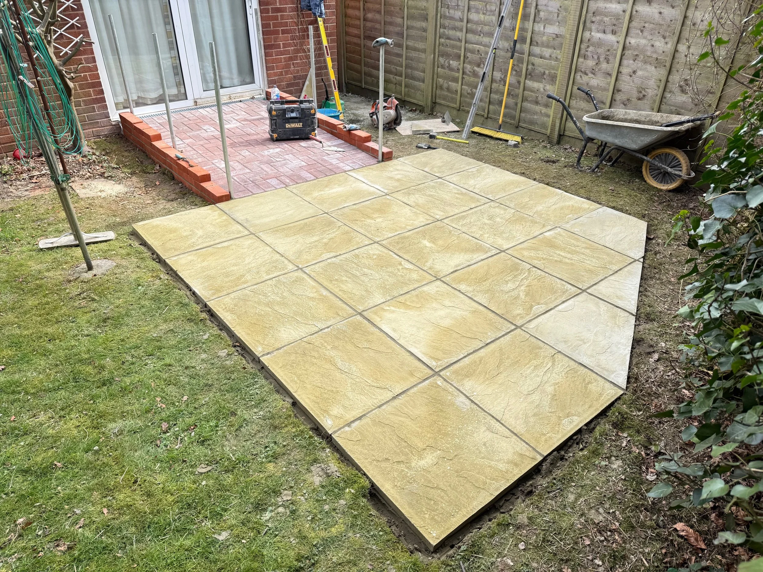 A backyard patio area under construction with freshly laid yellowish stone tiles, a brick platform with paving stones, and construction tools scattered around