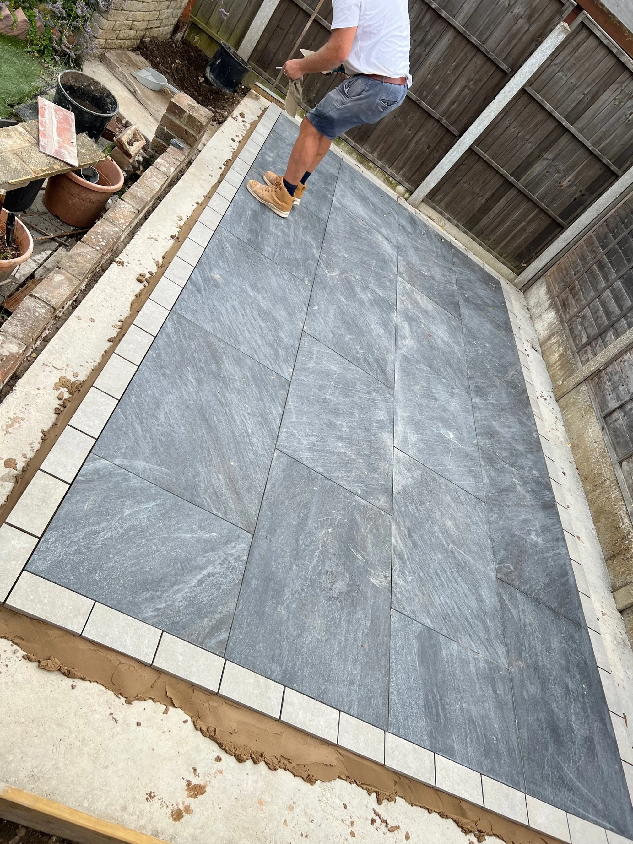 A person installing large gray paving stones in a backyard patio area, with a curb of smaller white tiles surrounding the patio. There are potted plants and construction tools along the edge.