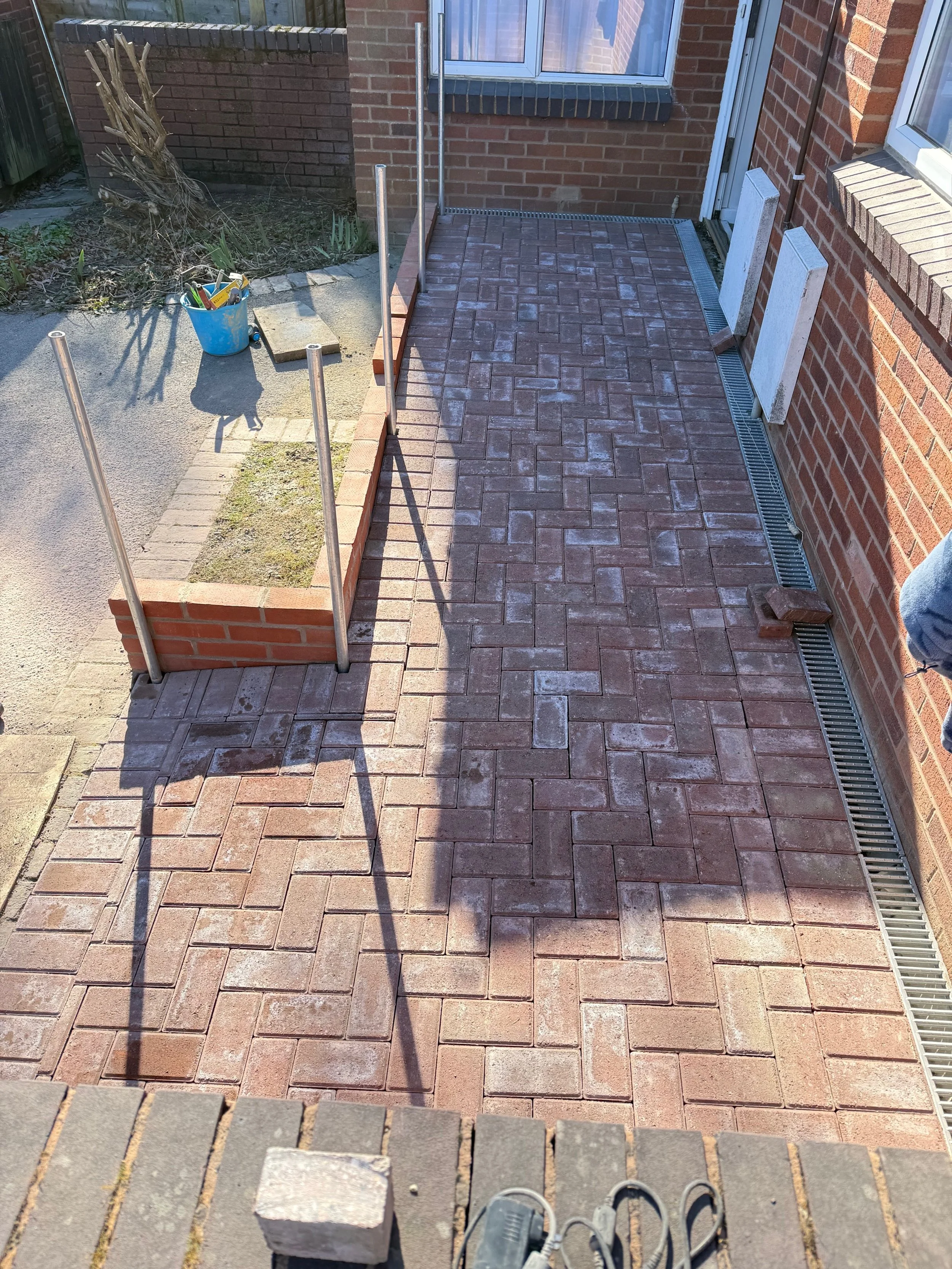A newly paved brick patio or walkway outside a brick house, partially enclosed with a metal railing, with construction tools and materials nearby.