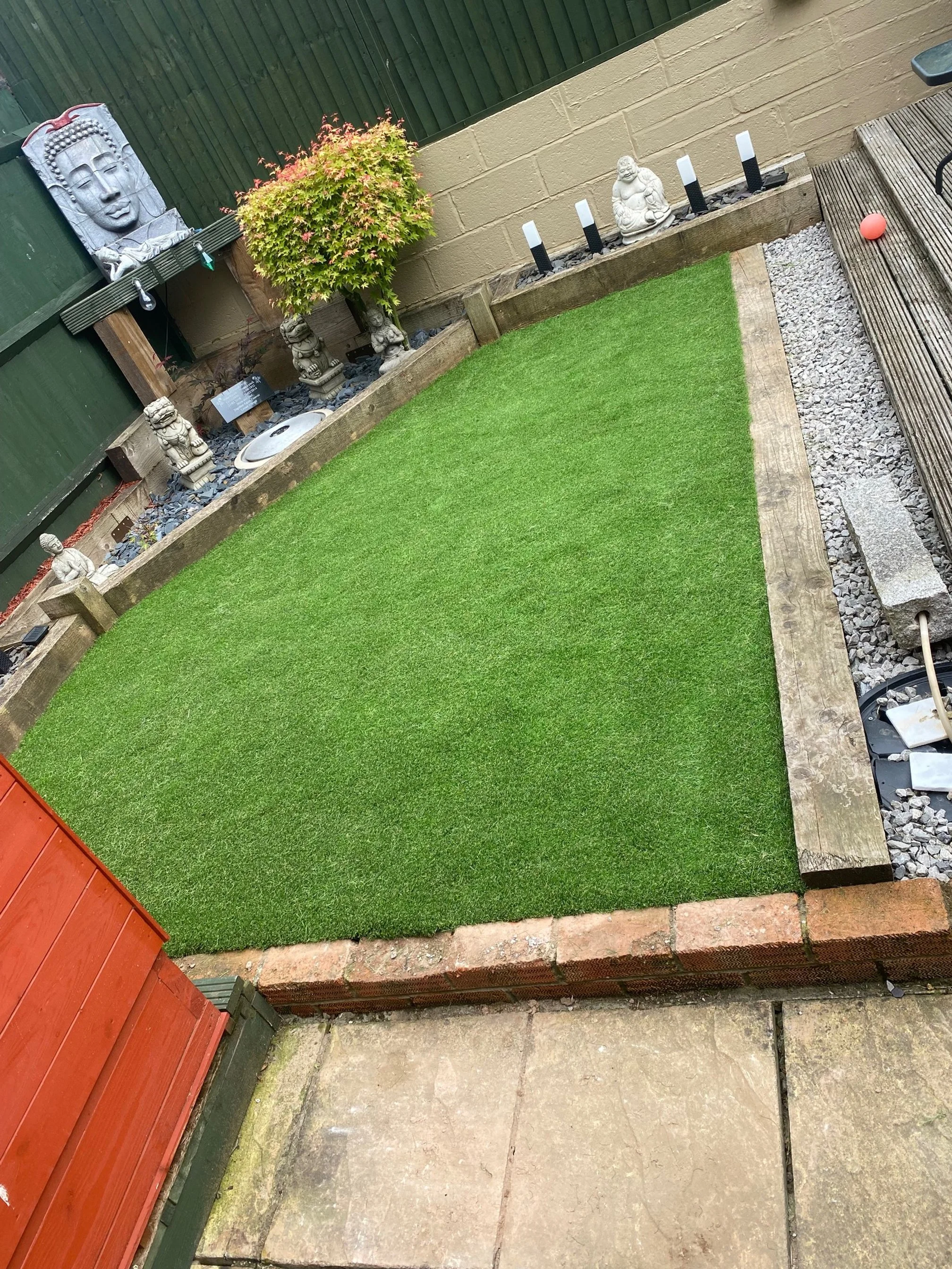 Backyard with artificial grass, decorative Buddha statues, small trees, candle-like light holders, a red ball, and a wooden deck.