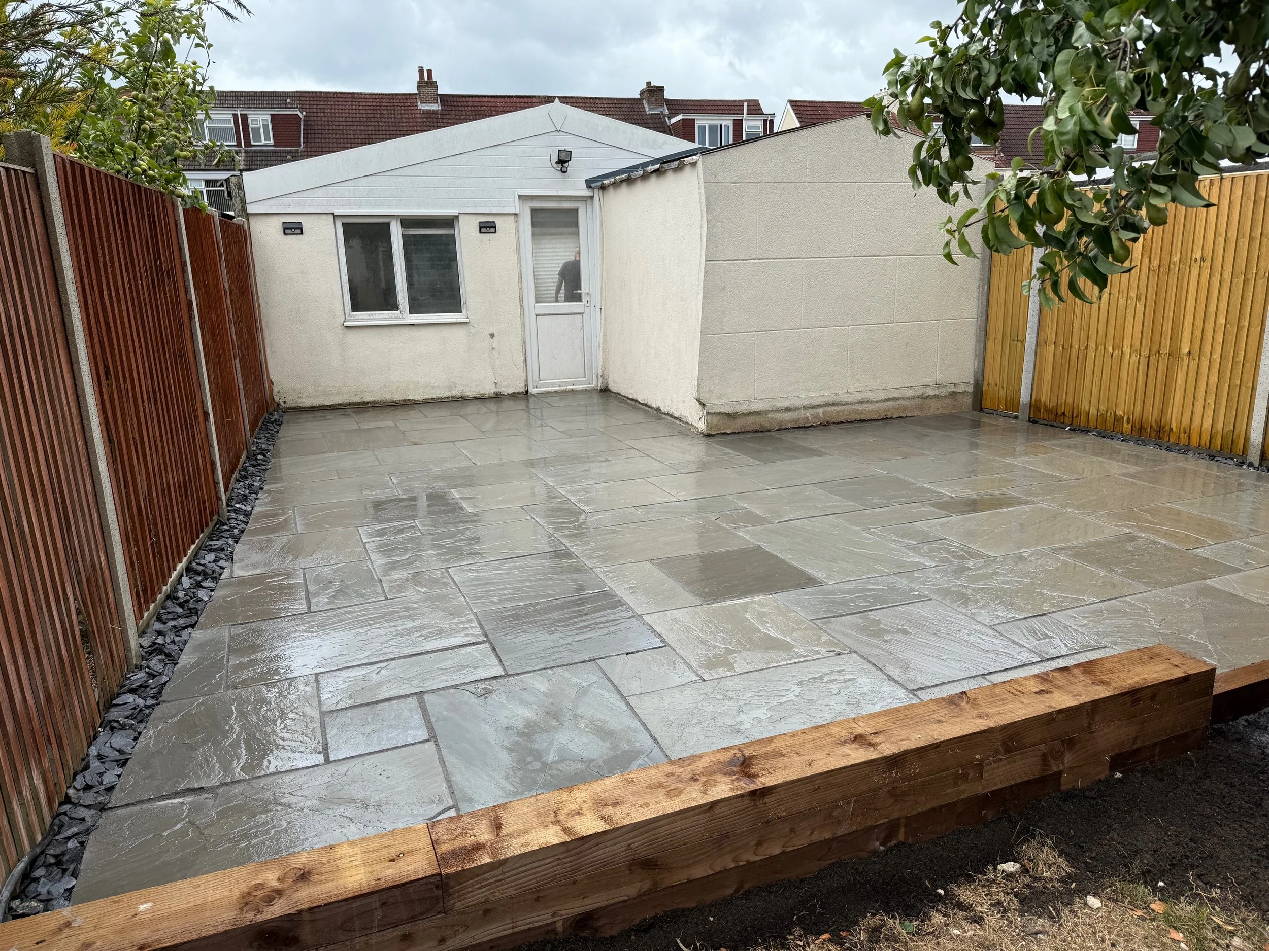 Finish of a newly paved outdoor patio area with light grey stone tiles, surrounded by wooden fencing and a small white garden shed.