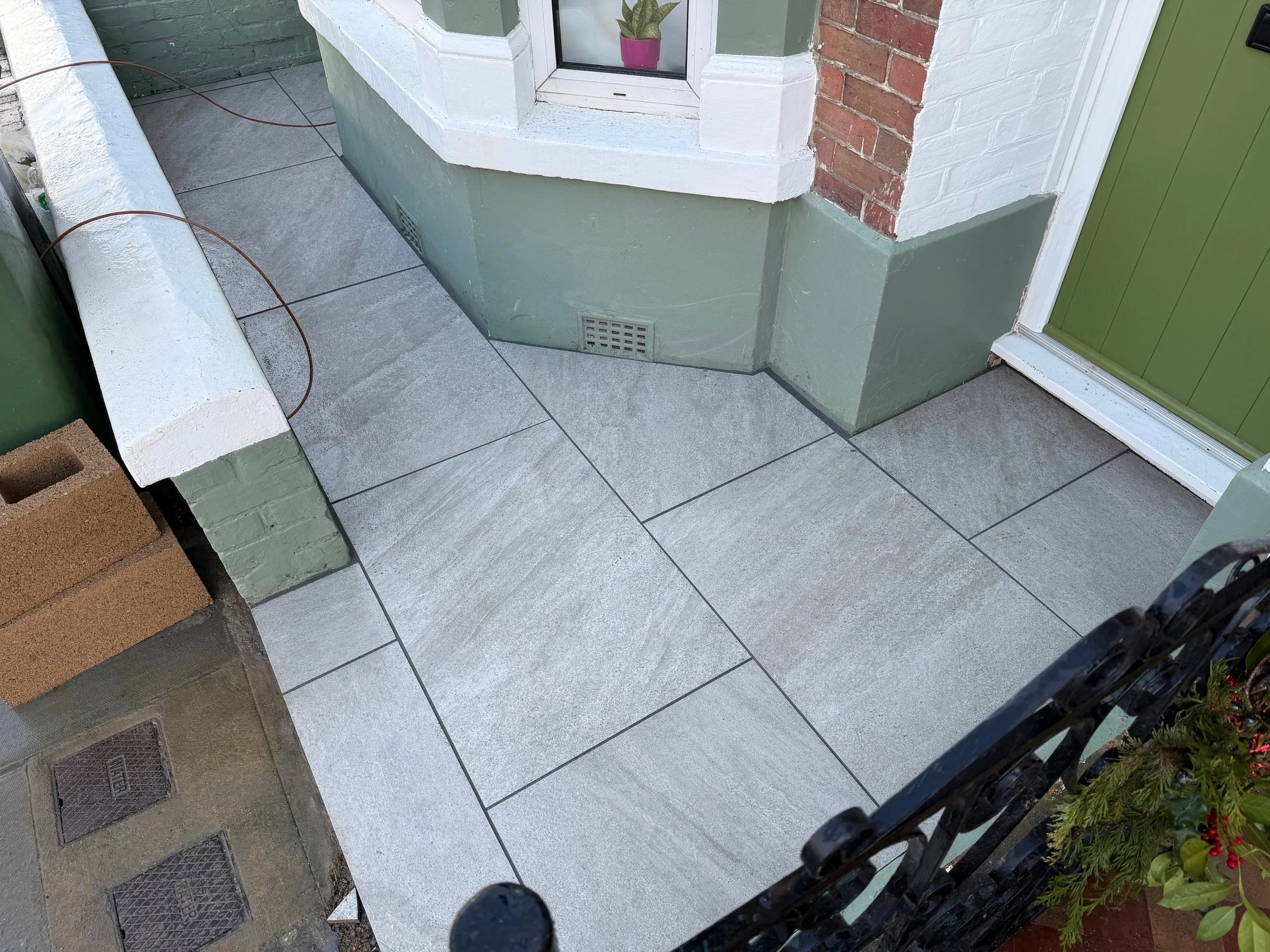 View of an outdoor entrance area featuring tiled flooring, a green door, a bay window, a brick wall, and a decorative black metal fence, with some potted plants visible.