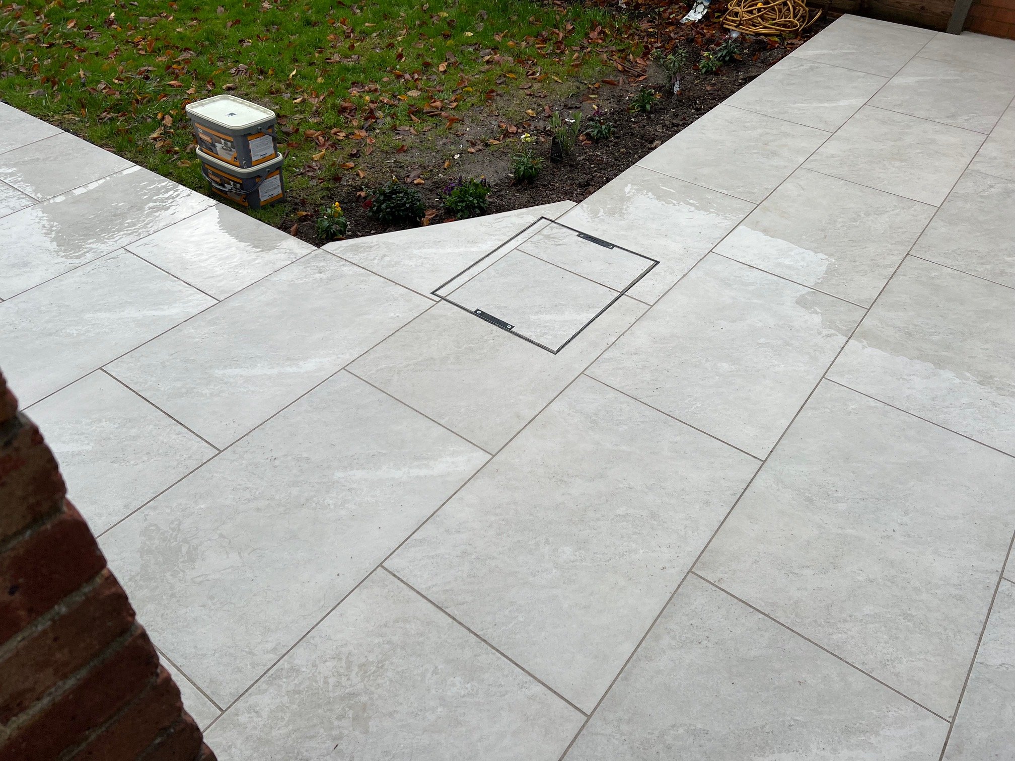 Outside patio with large beige tiles, garden bed with small plants and some fallen leaves, and a brick wall.